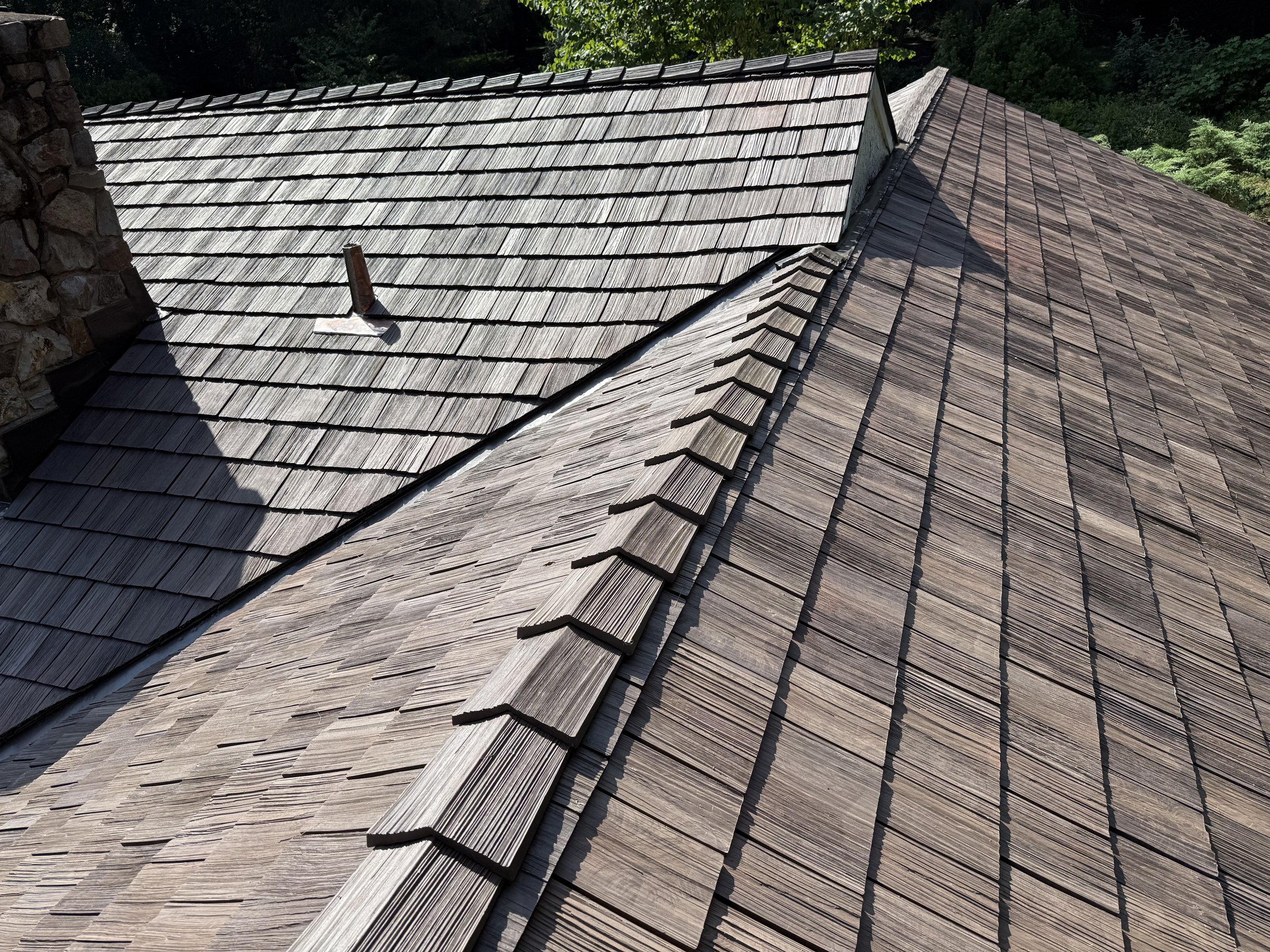 Close-up view of a house roof with overlapping wooden shingles in natural light, showing variations in color and texture, and a chimney with a pipe protruding from it.