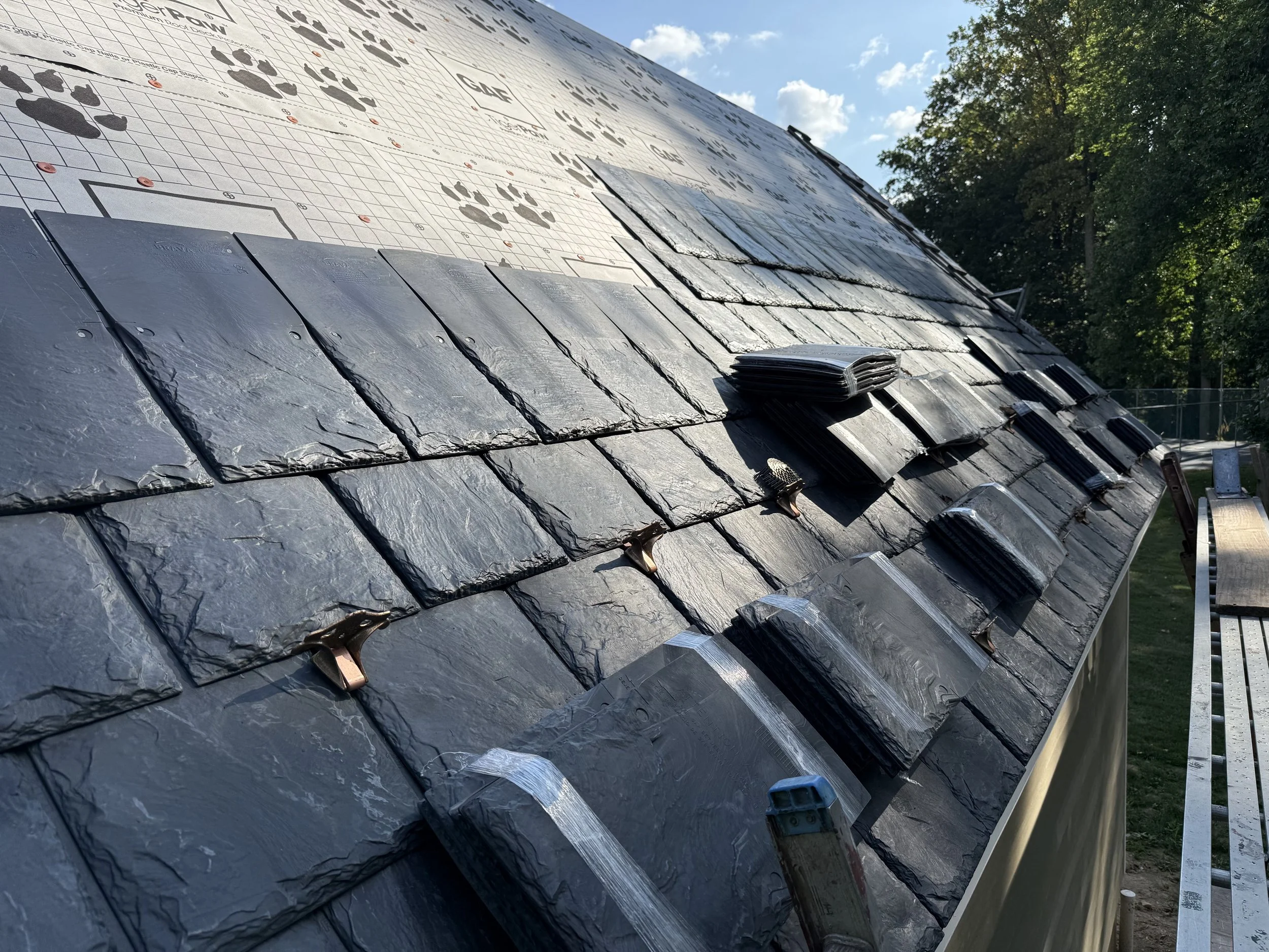 Close-up view of a roof under construction with slate tiles, clips, and roofing tools, with a background of trees and a sky with clouds.
