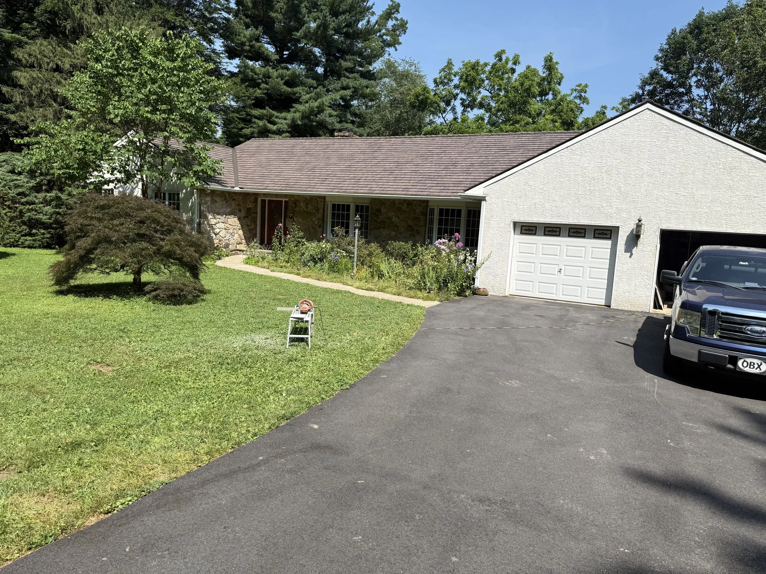 A white house with a brown roof and attached garage, driveway with a black truck, landscaped front yard with bushes and trees, and a small white stool with a garden hose on it.