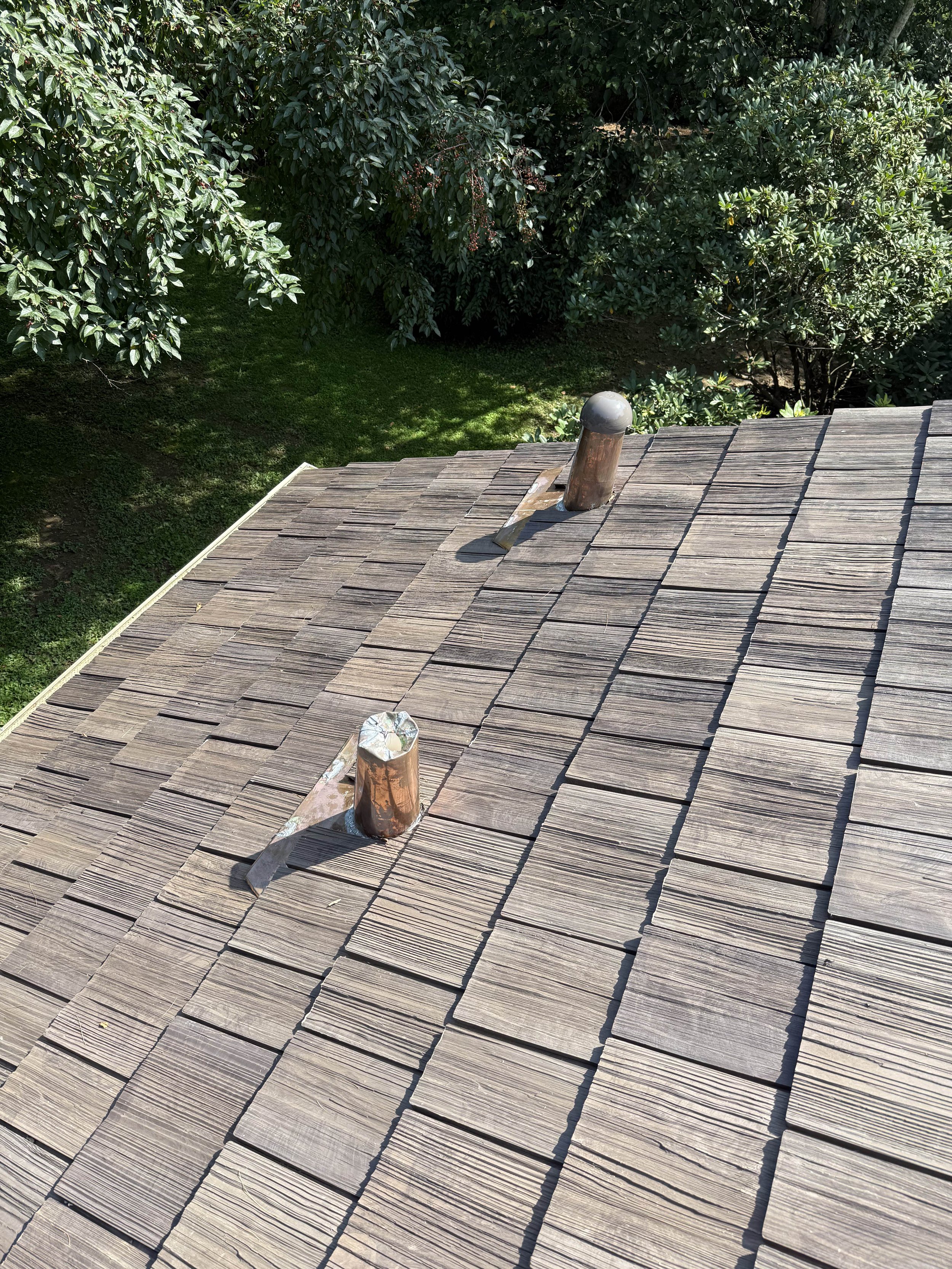 Top view of a wooden shingled roof with two metal roof vents, surrounded by green trees and grass.