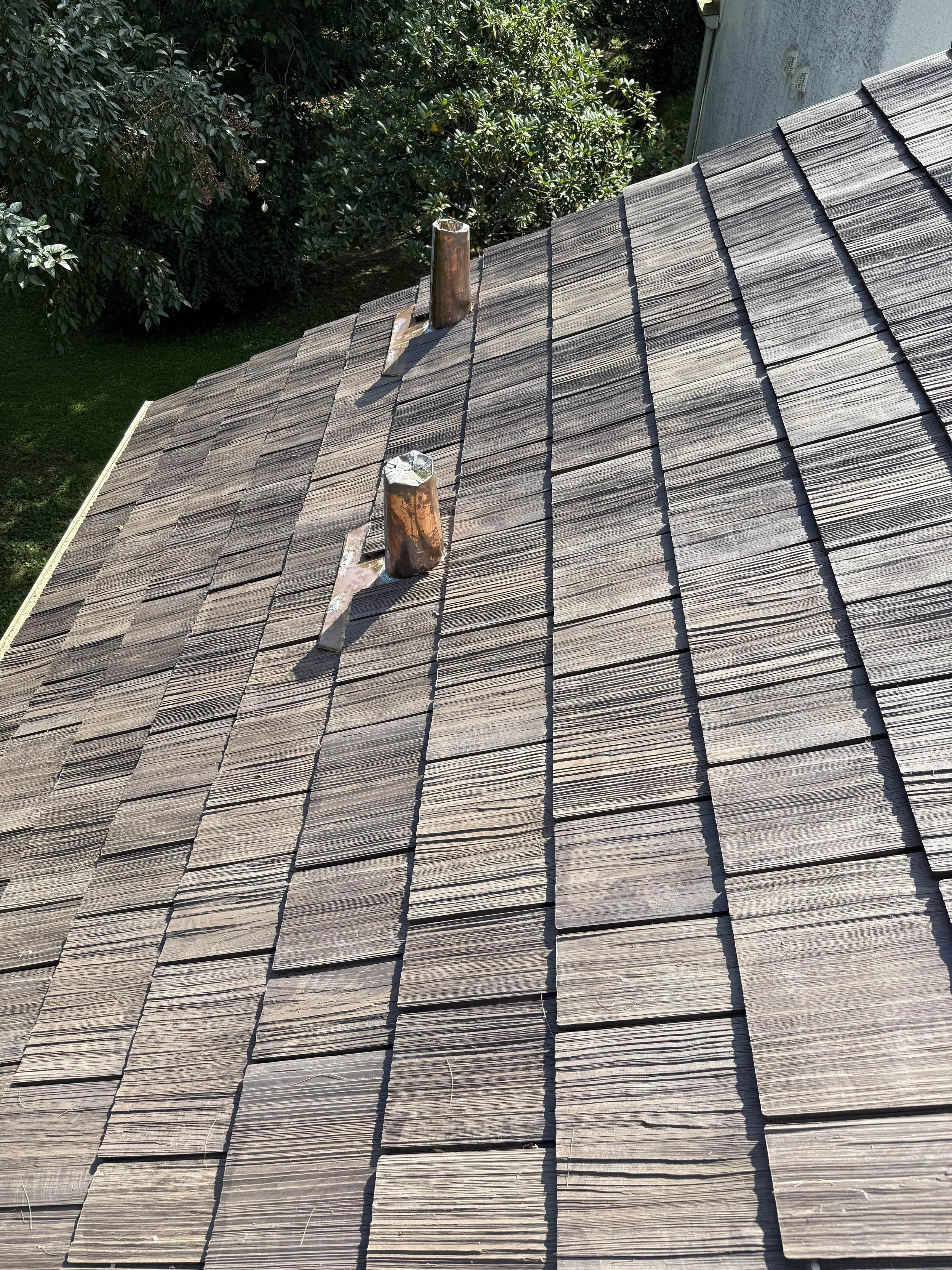 Wooden shingle roof with two vent pipes and surrounding green trees and part of a house wall.