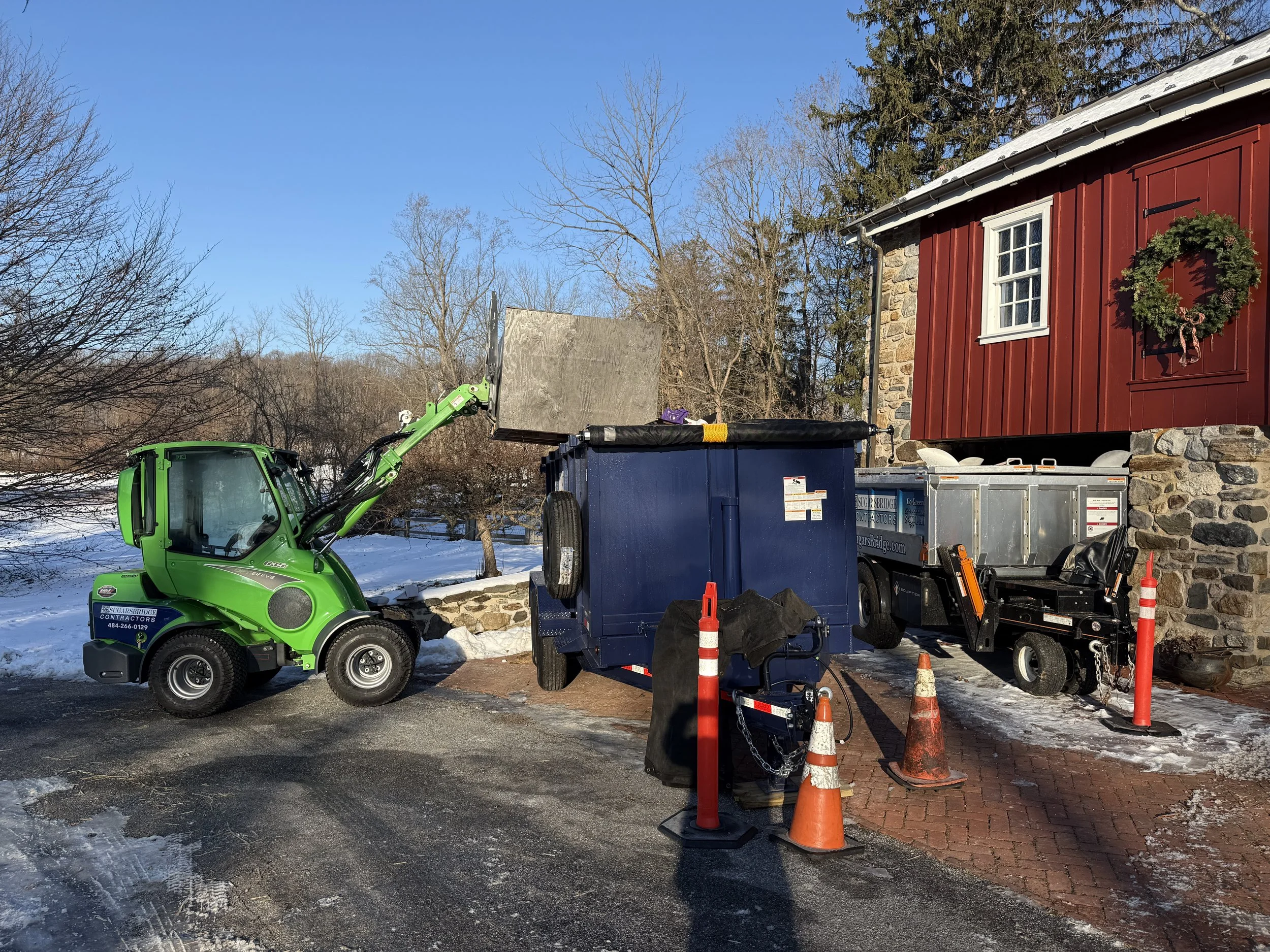 A green loader is pouring material into a blue dumpster outside a red building with a holiday wreath, snow and ice on the ground.
