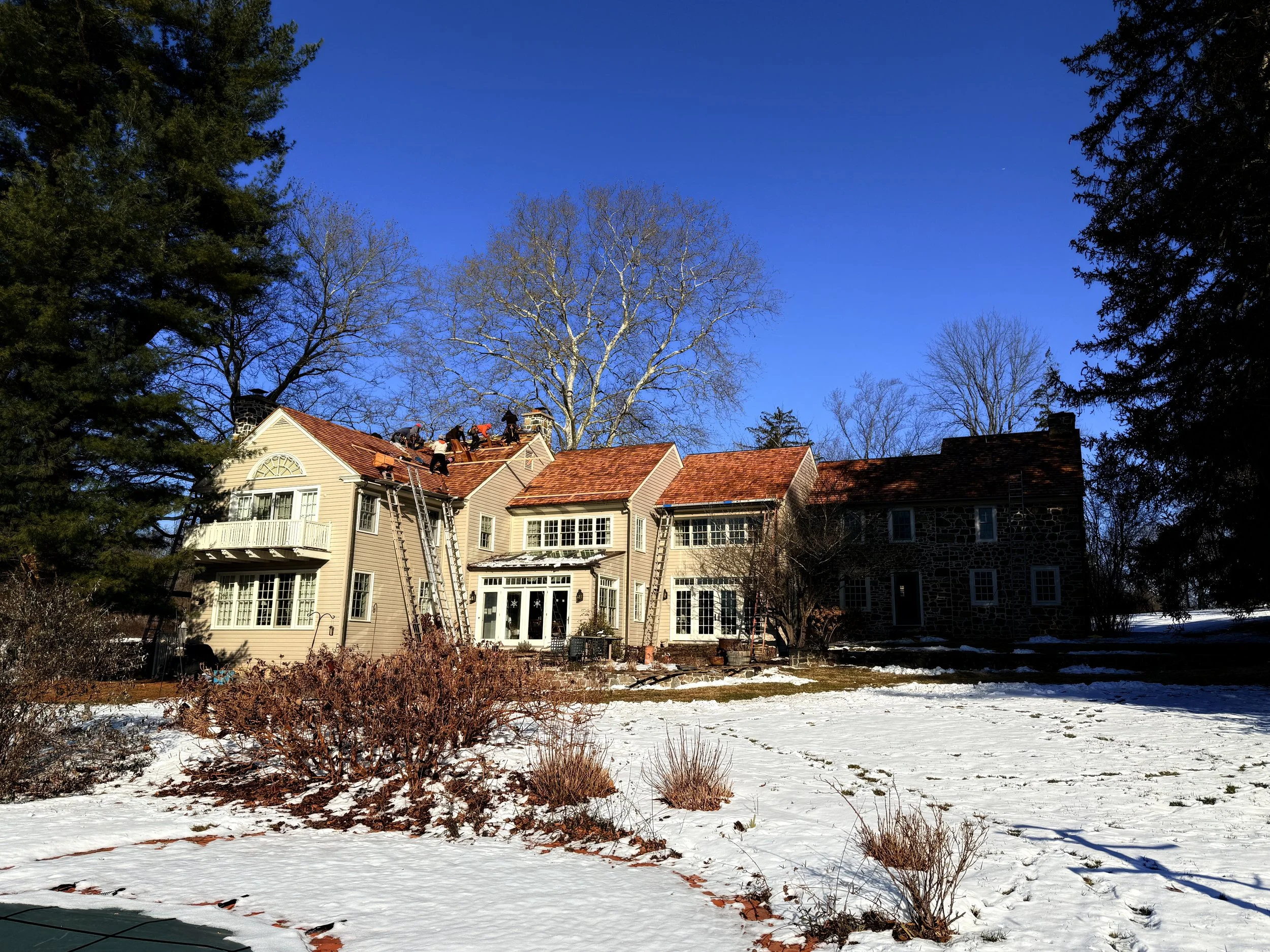 A house with workers on the roof, surrounded by snow and leafless trees on a clear, sunny day.