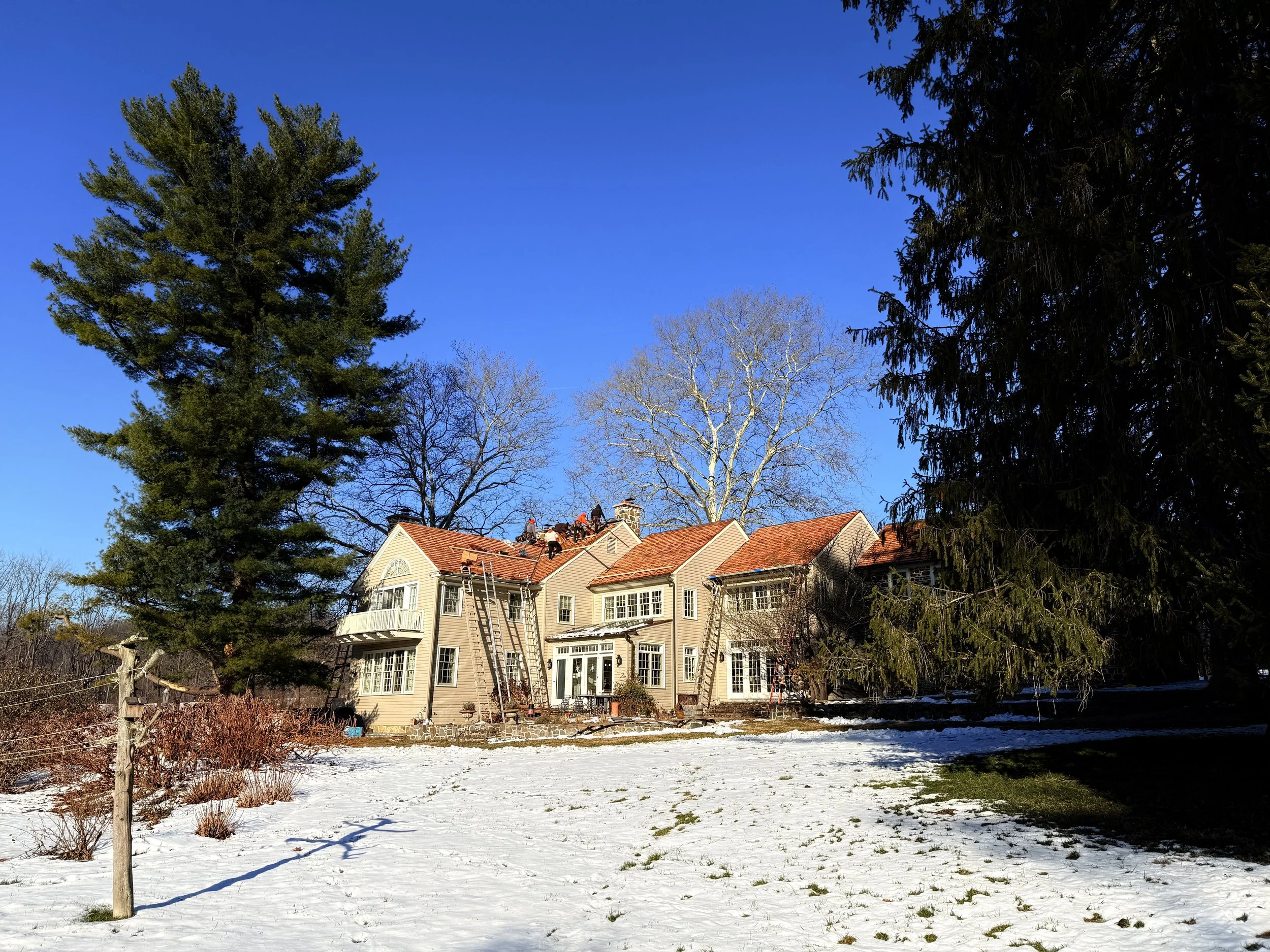 A historic Chester County house with beige siding and red roof tiles is being maintained on a bright winter day with snow on the ground. Tall trees surround the house, some with bare branches and others with green foliage, under a clear blue sky.