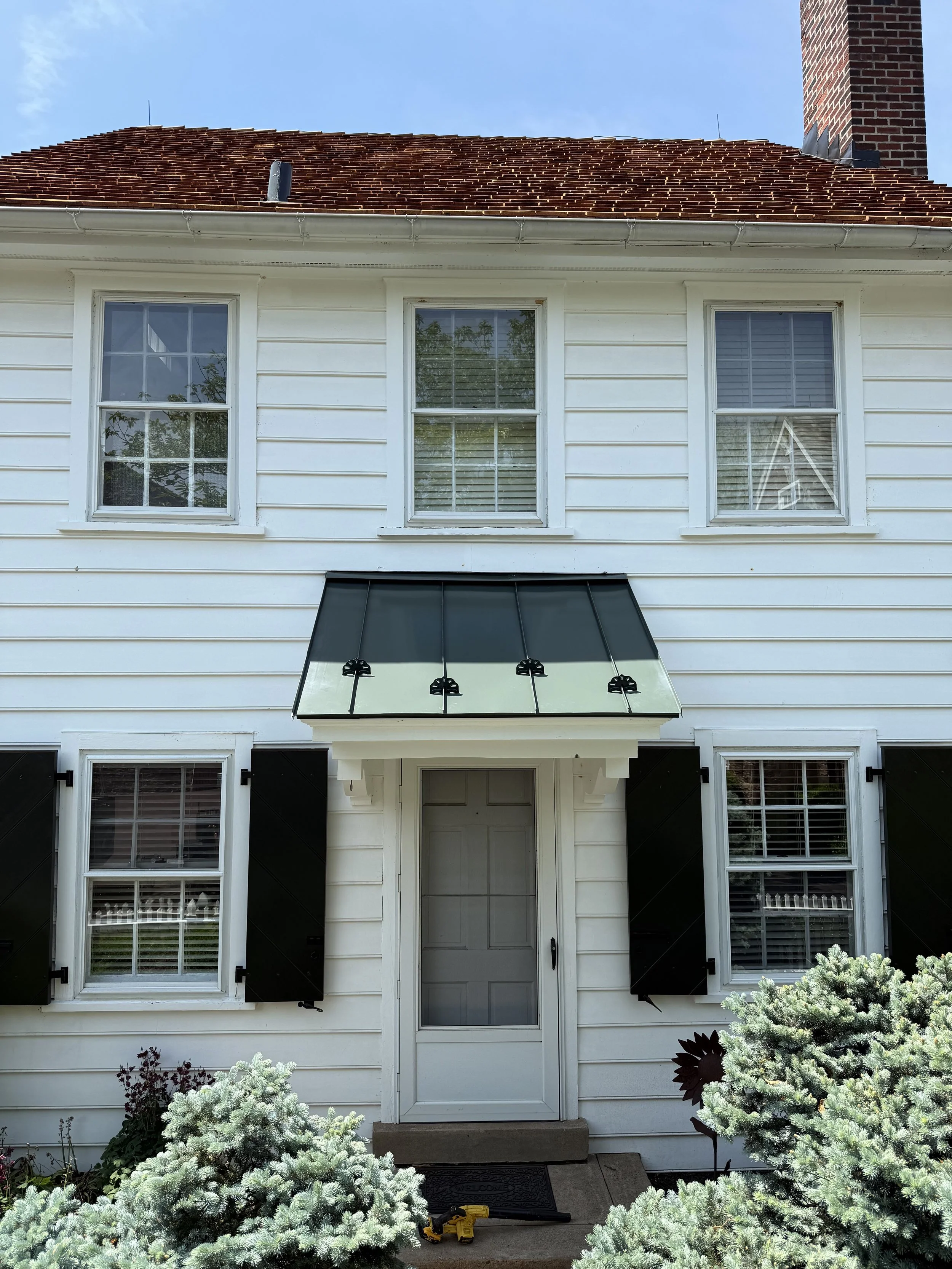 Front view of a white two-story house with three windows on the upper floor, black shutters, and a door with a glass pane on the lower level. A black awning over the door, decorative bushes in front, and tools on the porch.