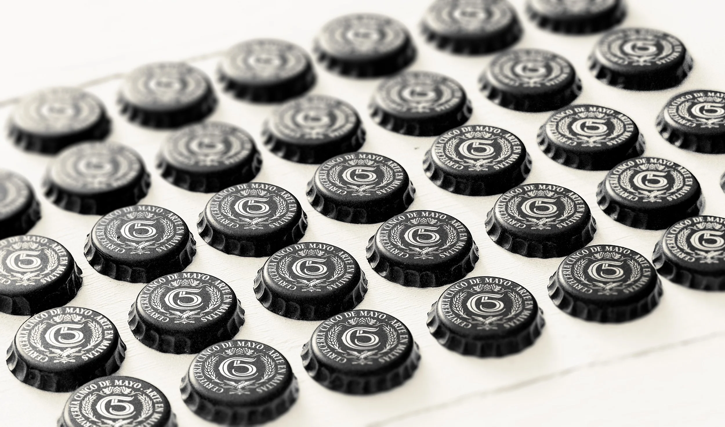 Multiple black bottle caps arranged in rows, each featuring the Cervecería Cinco de Mayo logo with the “C5” monogram, barley, hops, and the phrase “Arte en Maltas”; showcasing consistent branding, premium Mexican craft beer identity, and attention to