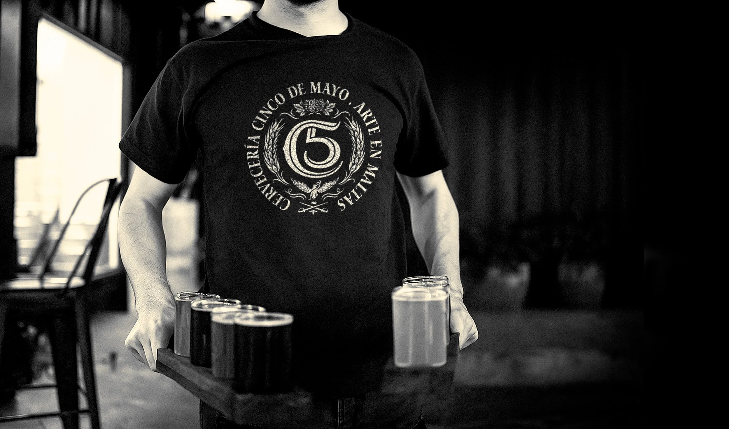 Server wearing a black Cervecería Cinco de Mayo t-shirt with the “C5” monogram and “Arte en Maltas” logo, holding a wooden tray with a craft beer flight; showcasing branded apparel, premium Mexican brewery identity, and artisanal tasting experience i