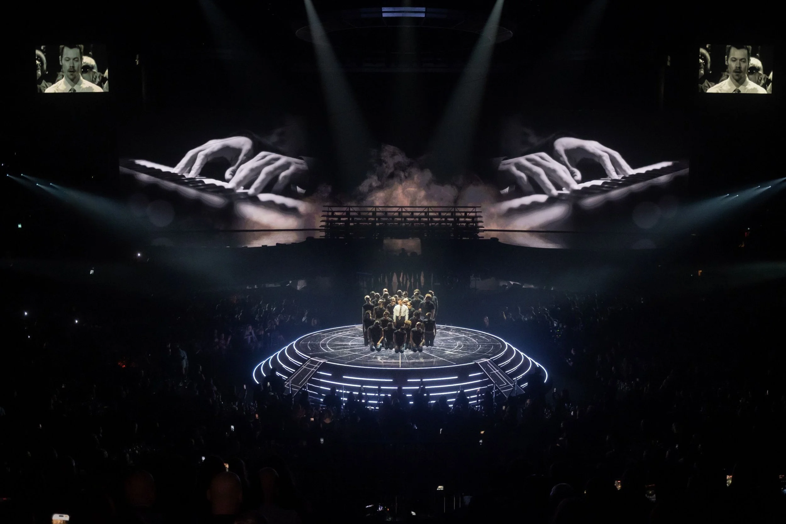 Picture of performance of singer Harry Style surrounded by multiple dancers on lit lower stage background screens displaying a pianist hands playing the instrument