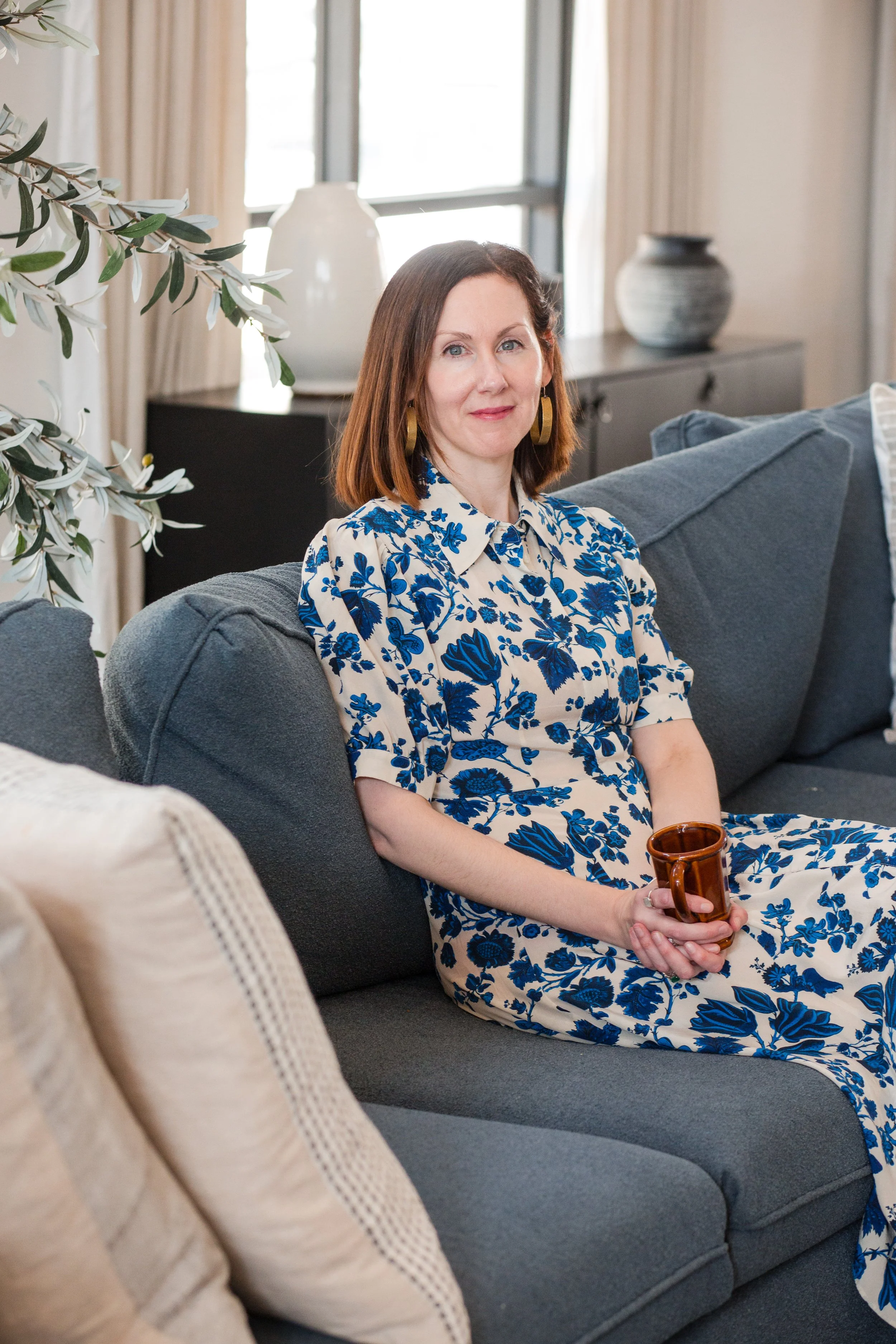 Woman with shoulder-length brown hair wearing a blue and beige floral dress, sitting on a gray sofa with a brown mug in her hand, in a brightly lit living room.