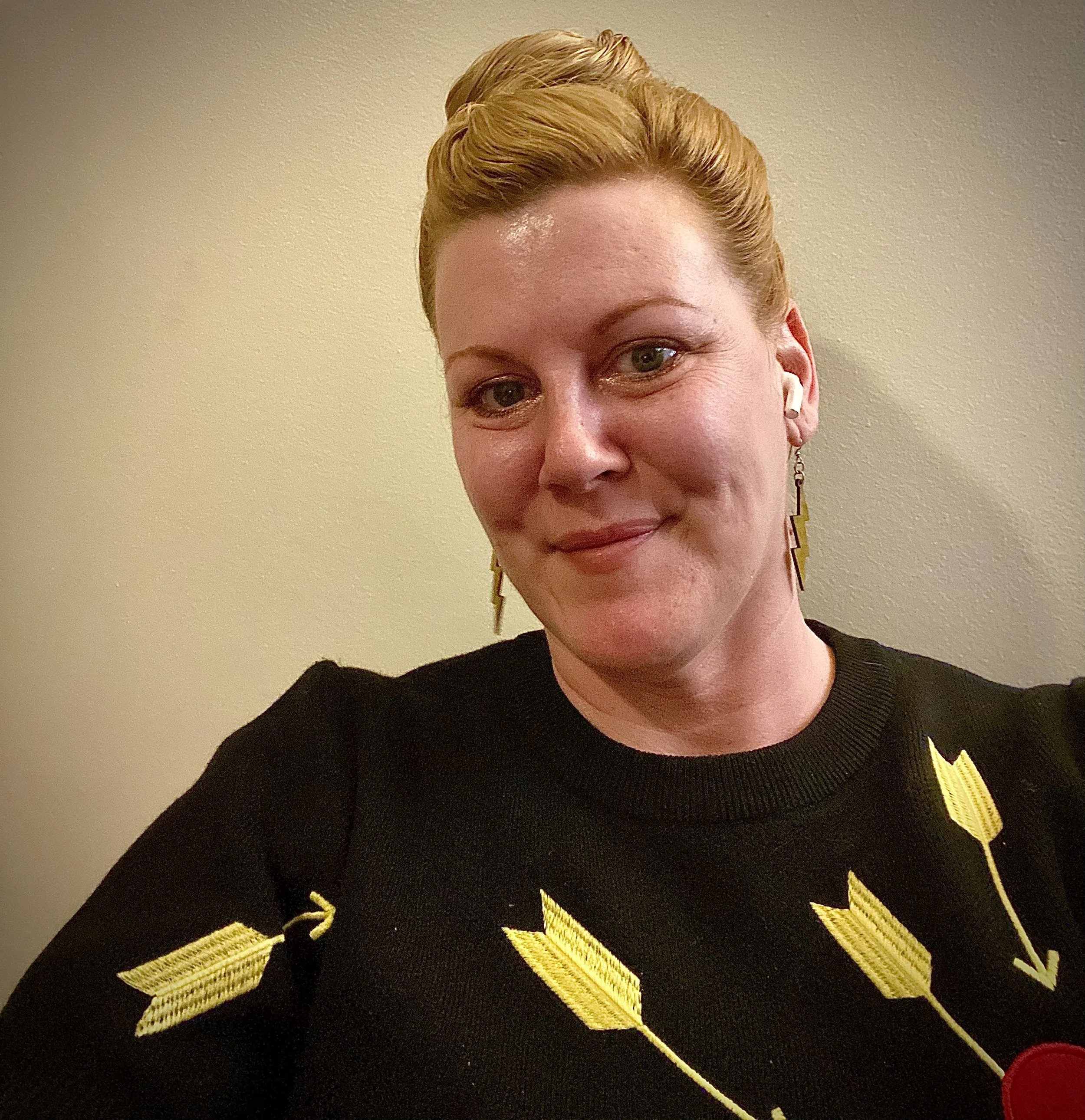 A woman with blonde hair styled in an updo, wearing a black sweater with yellow arrow designs, and gold lightning bolt earrings, standing against a plain beige wall.