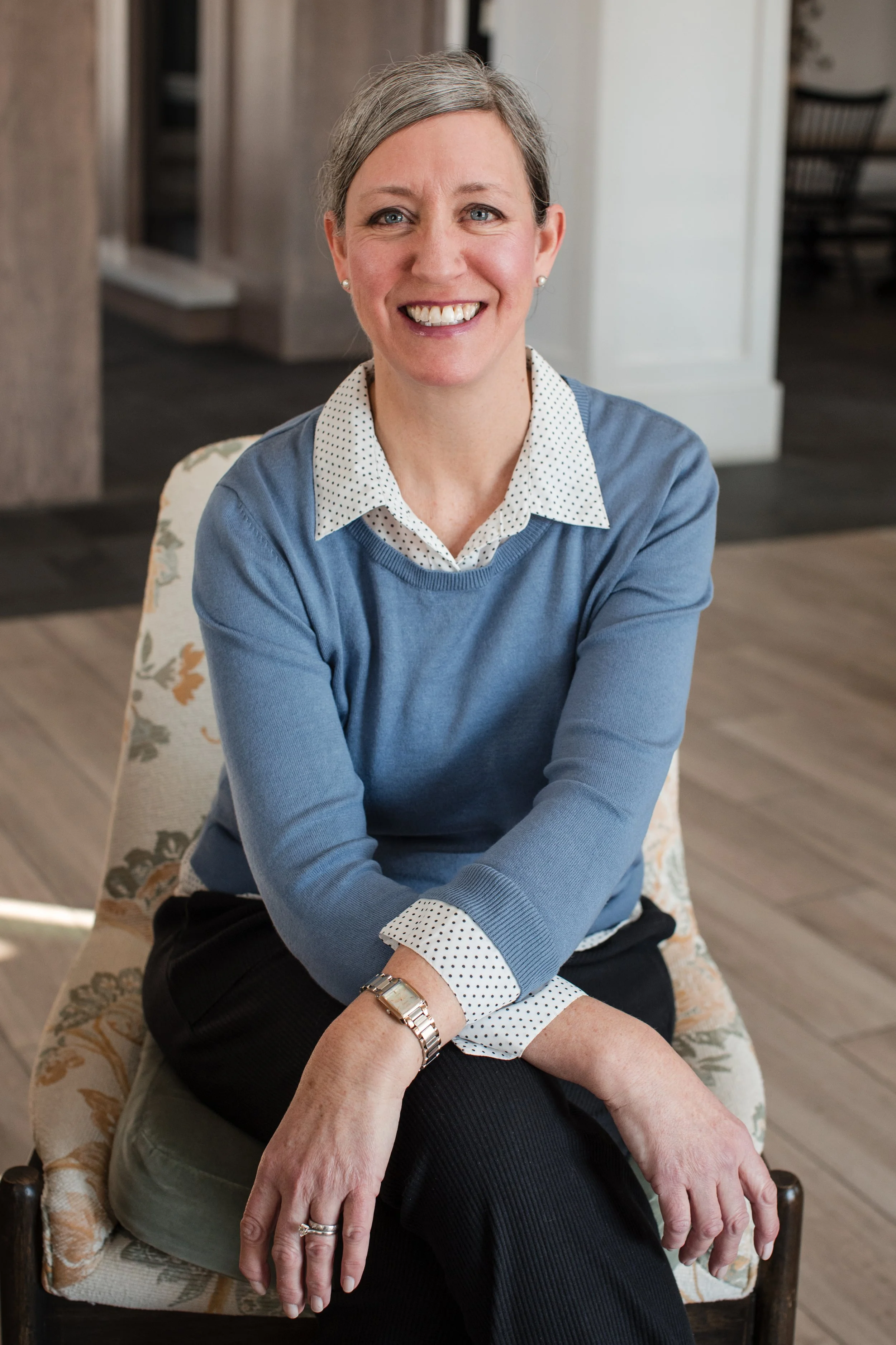 A woman with short gray hair and blue eyes, smiling while sitting on a patterned chair in an indoor setting. She is wearing a light blue sweater over a white blouse with black polka dots, black pants, and a wristwatch.