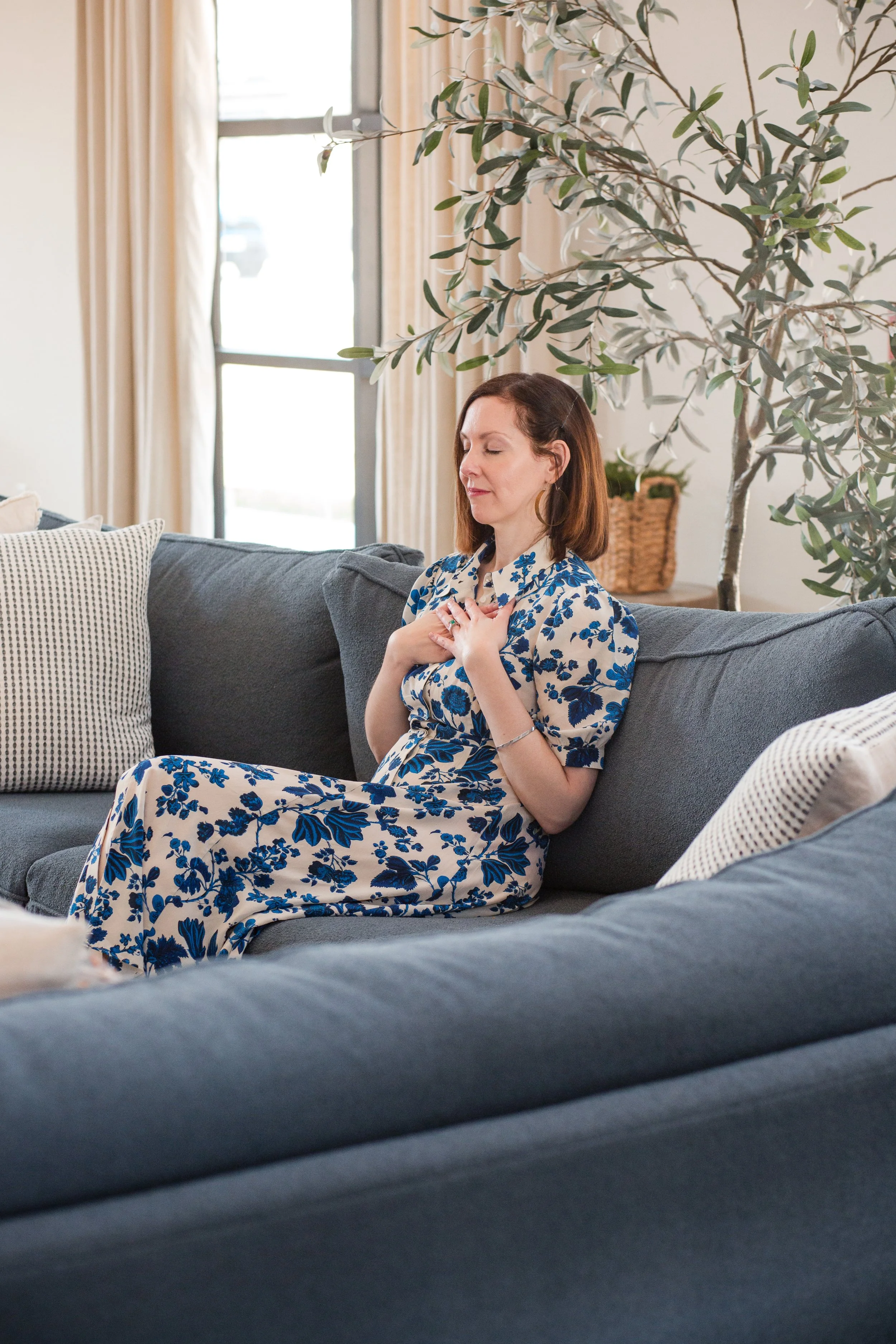A woman in a blue and white floral dress sits with her eyes closed and hands on her chest on a gray sofa in a bright living room with a large window, beige curtains, and a potted tree.