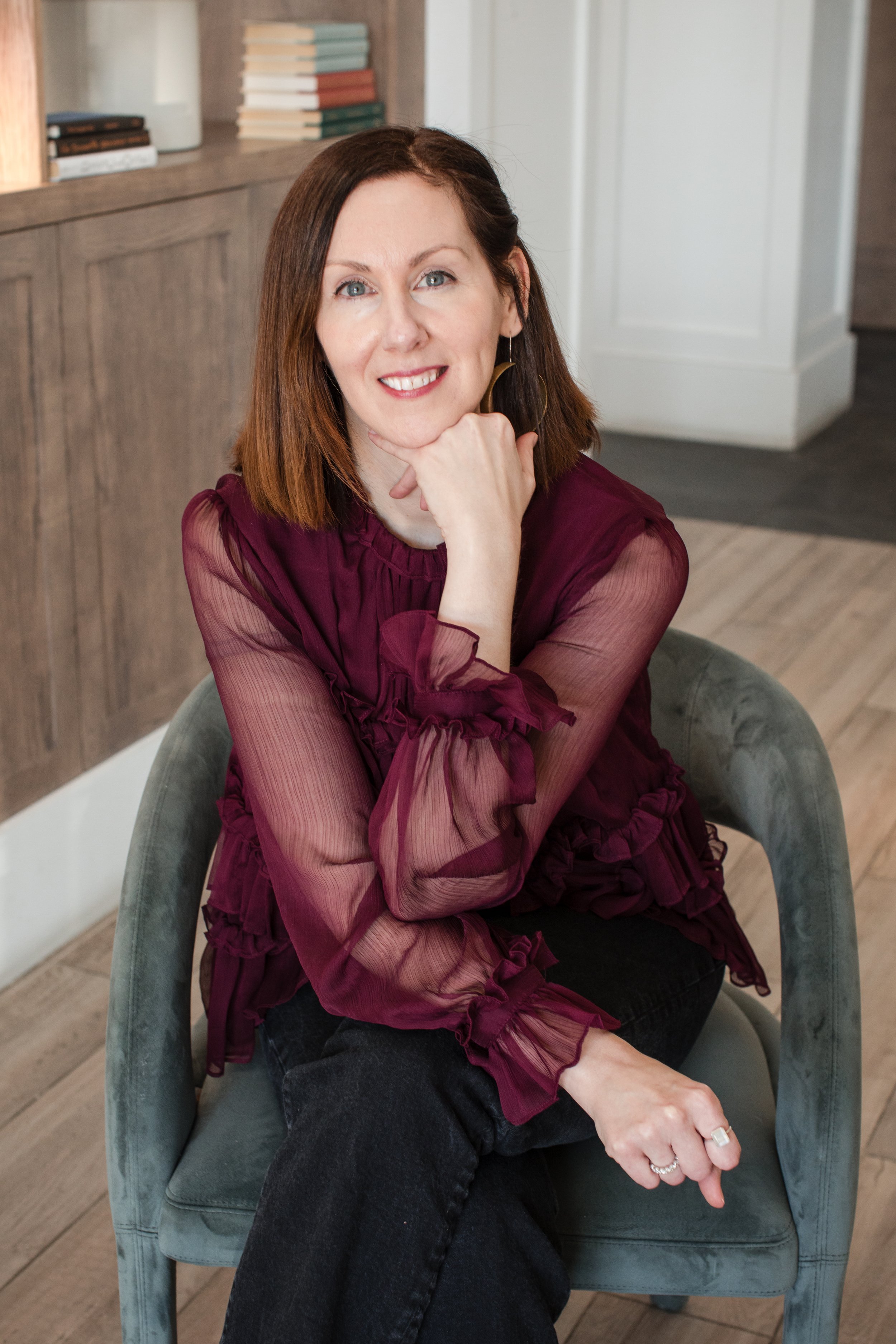 A woman with shoulder-length red hair sitting on a grey velvet armchair, wearing a sheer burgundy blouse with ruffles and black pants, smiling and resting her chin on her hand.