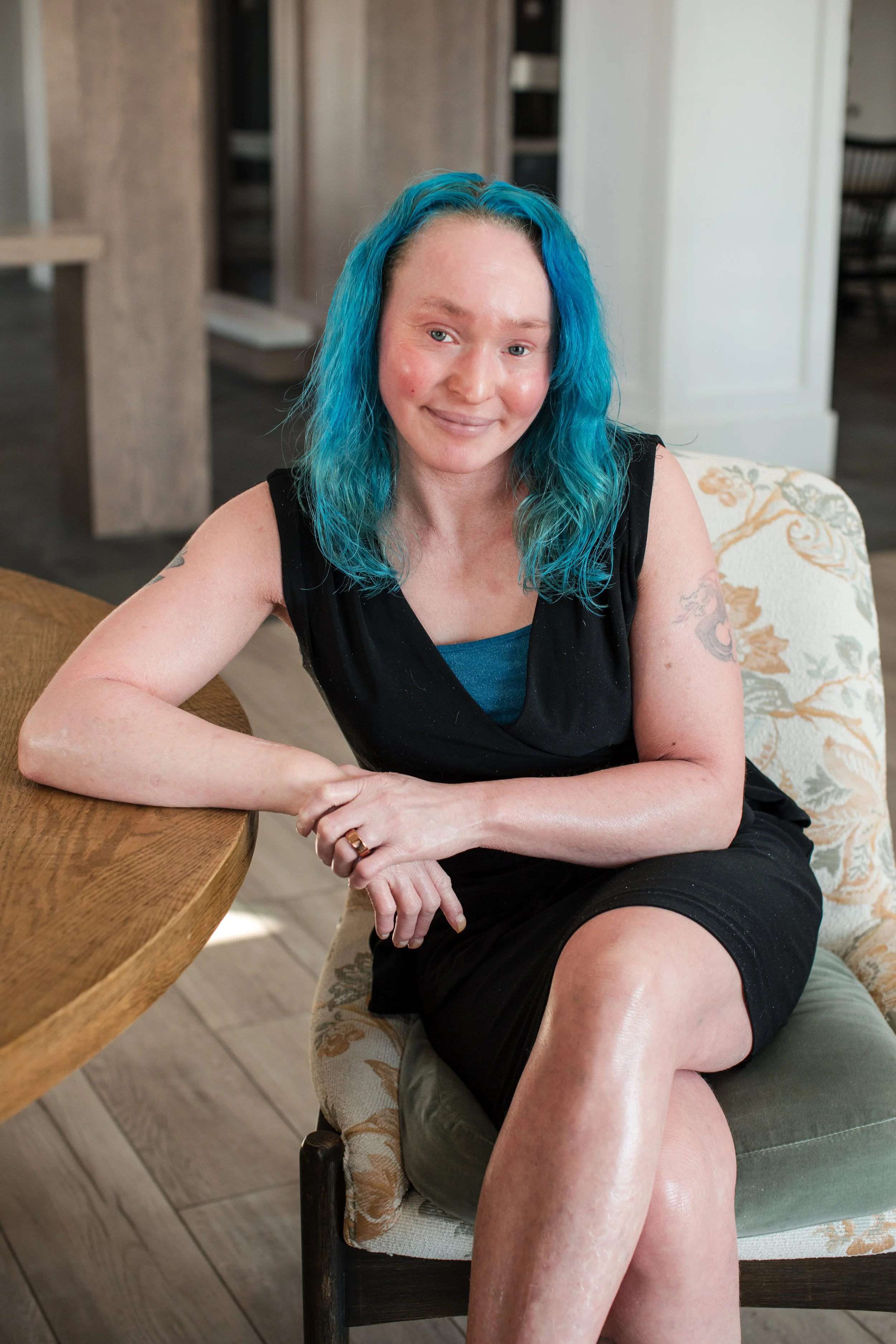A woman with bright blue hair sitting on an upholstered armchair, smiling softly, wearing a black dress, with tattoos on her arms, in a cozy indoor setting.
