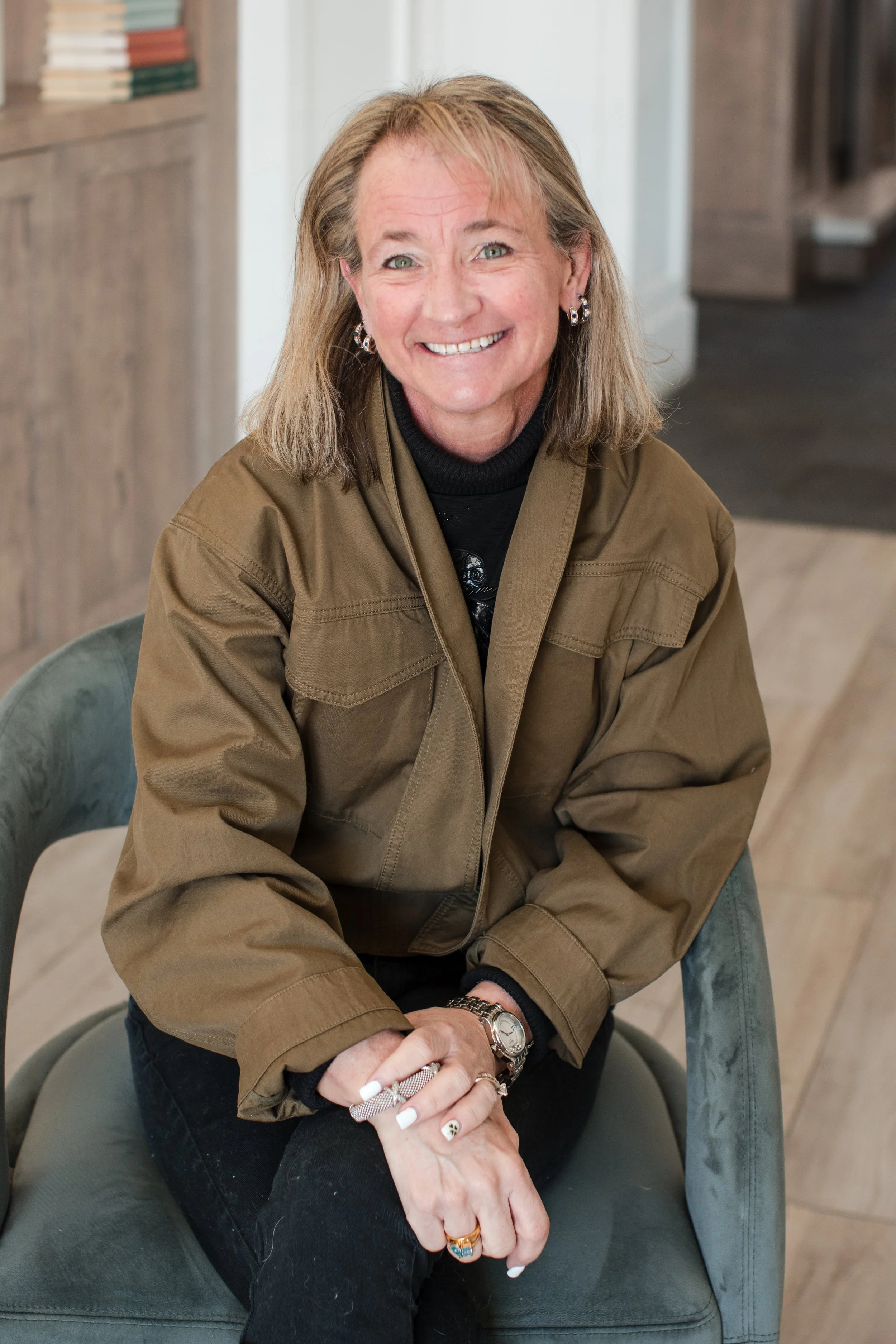 A woman with shoulder-length blonde hair smiling, sitting on a green upholstered chair indoors, wearing a brown jacket, black turtleneck, and jewelry.