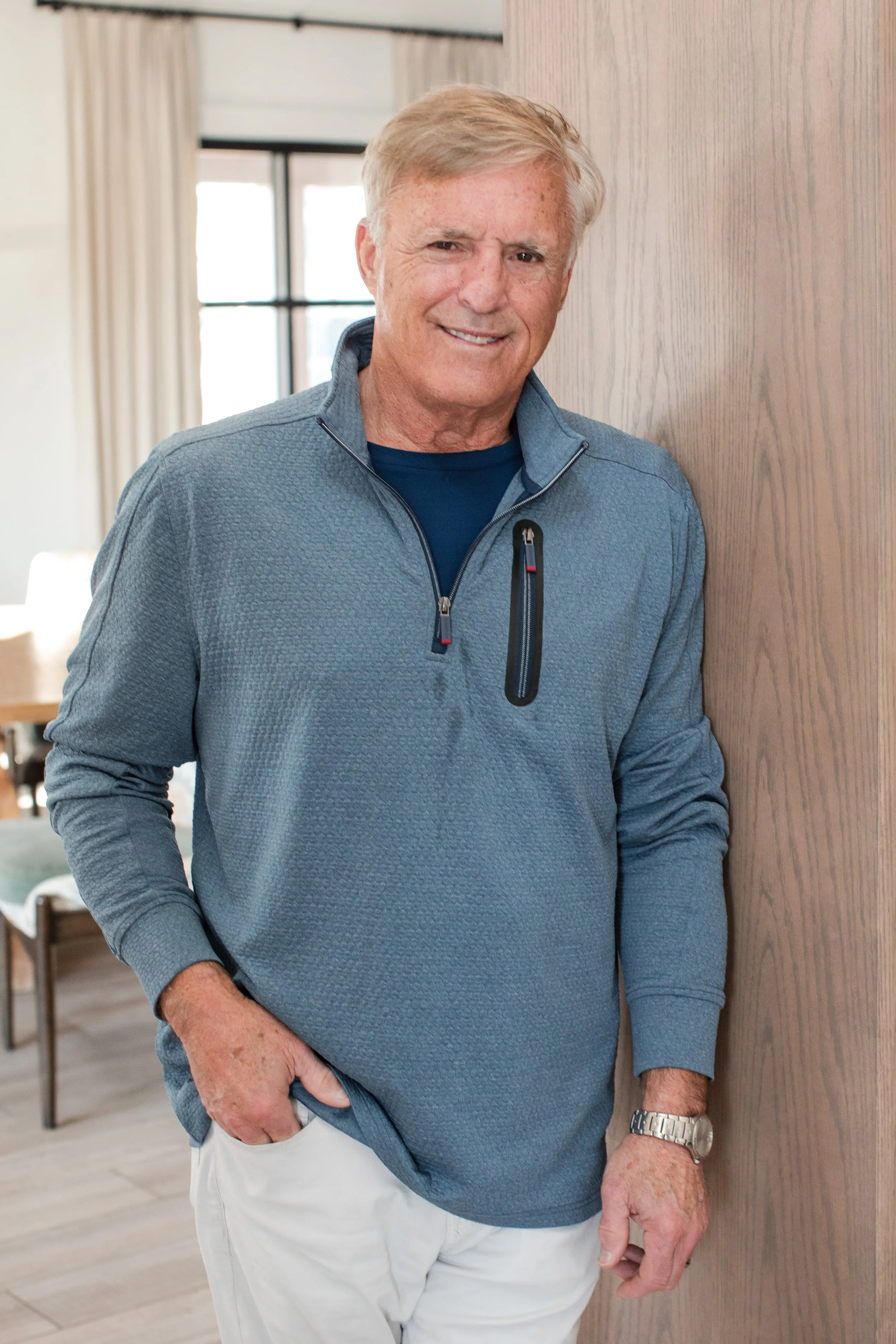 A smiling older man with light-colored hair, wearing a blue quarter-zip pullover, standing indoors against a wooden wall, with a background of home furniture and windows.