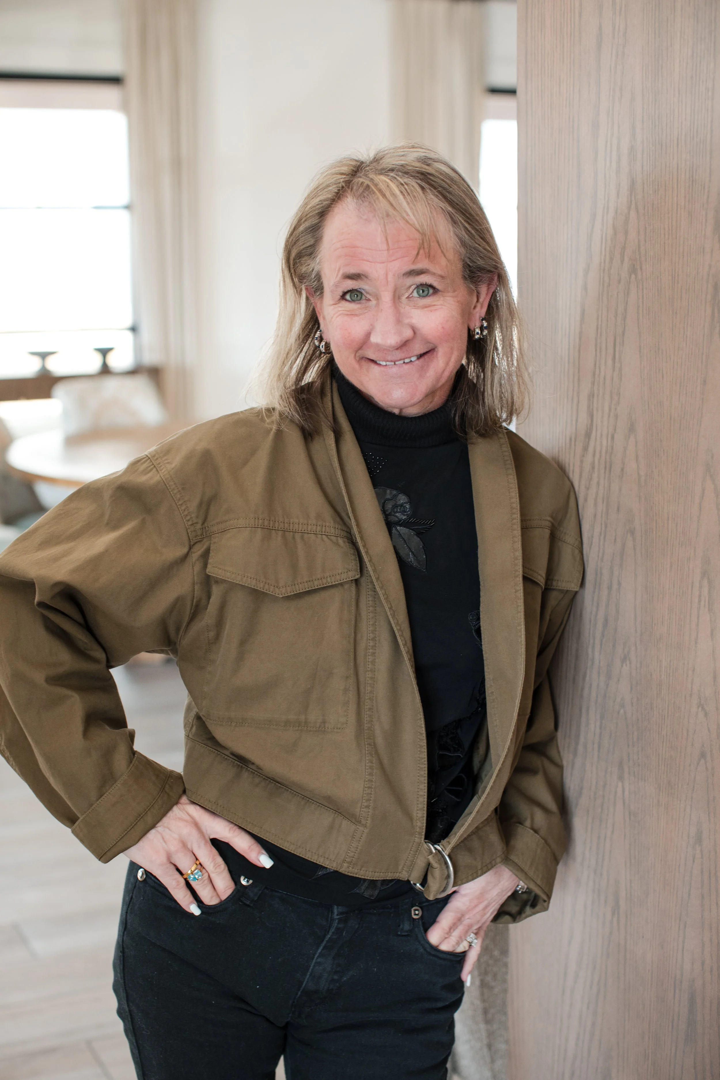A woman with shoulder-length blonde hair, wearing a black turtleneck and a brown jacket, standing near a wooden wall, smiling with a hand on her hip in a well-lit room with natural light and a dining area in the background.