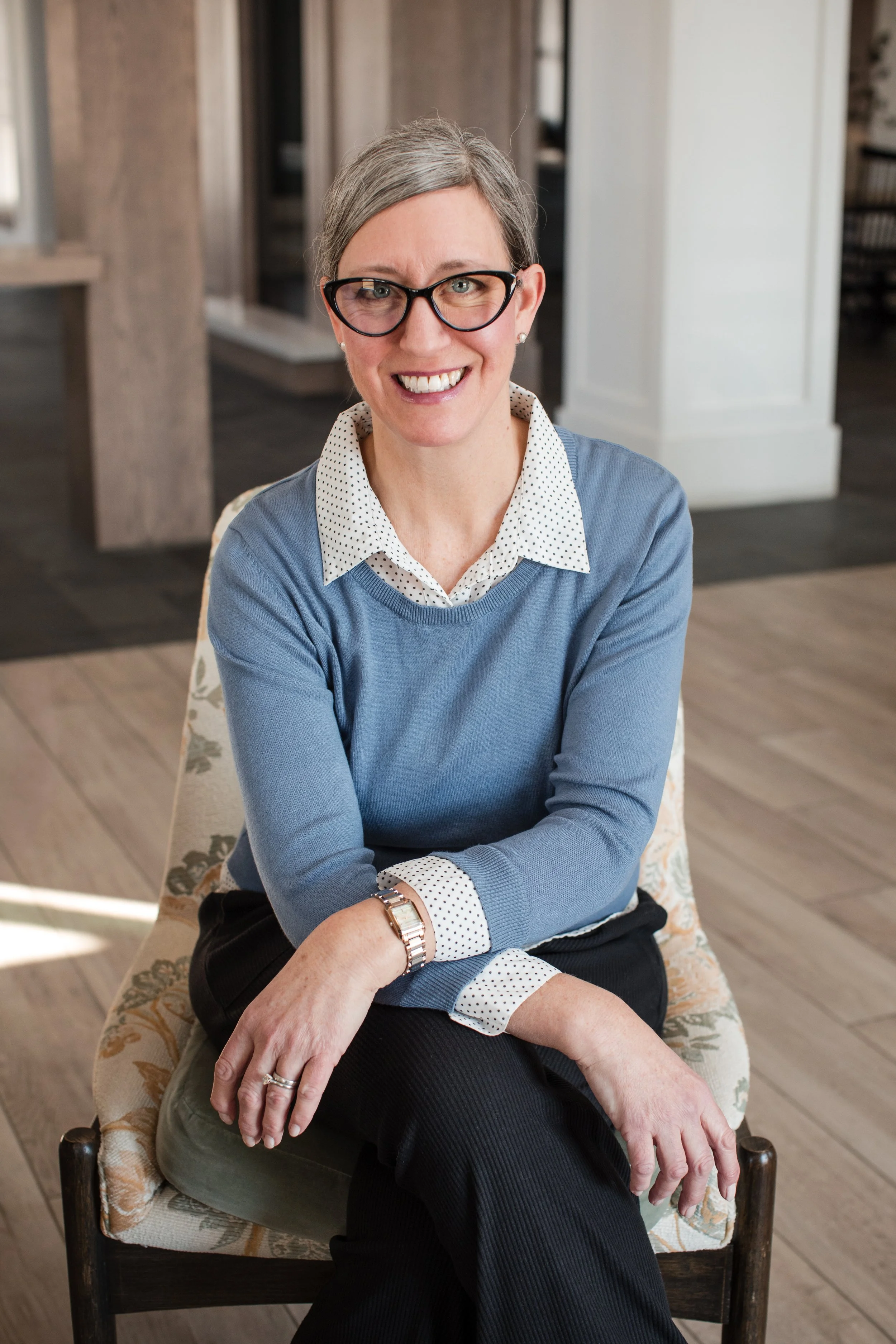 A smiling woman with gray hair, glasses, and a polka-dot collared shirt under a blue sweater, sitting on a patterned chair in a modern indoor setting.