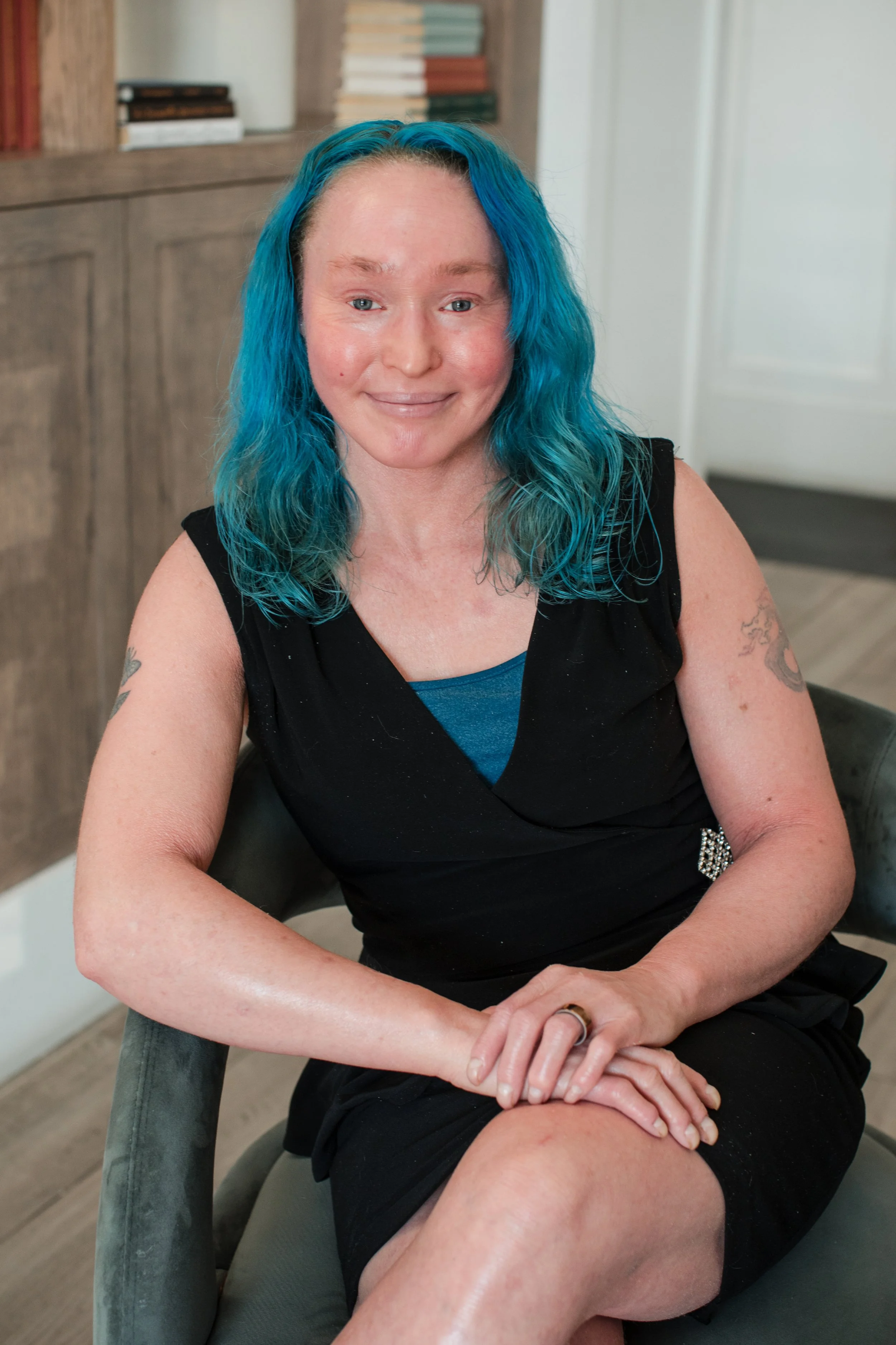A woman with blue wavy hair sitting on a chair, smiling at the camera, wearing a black dress and a ring.
