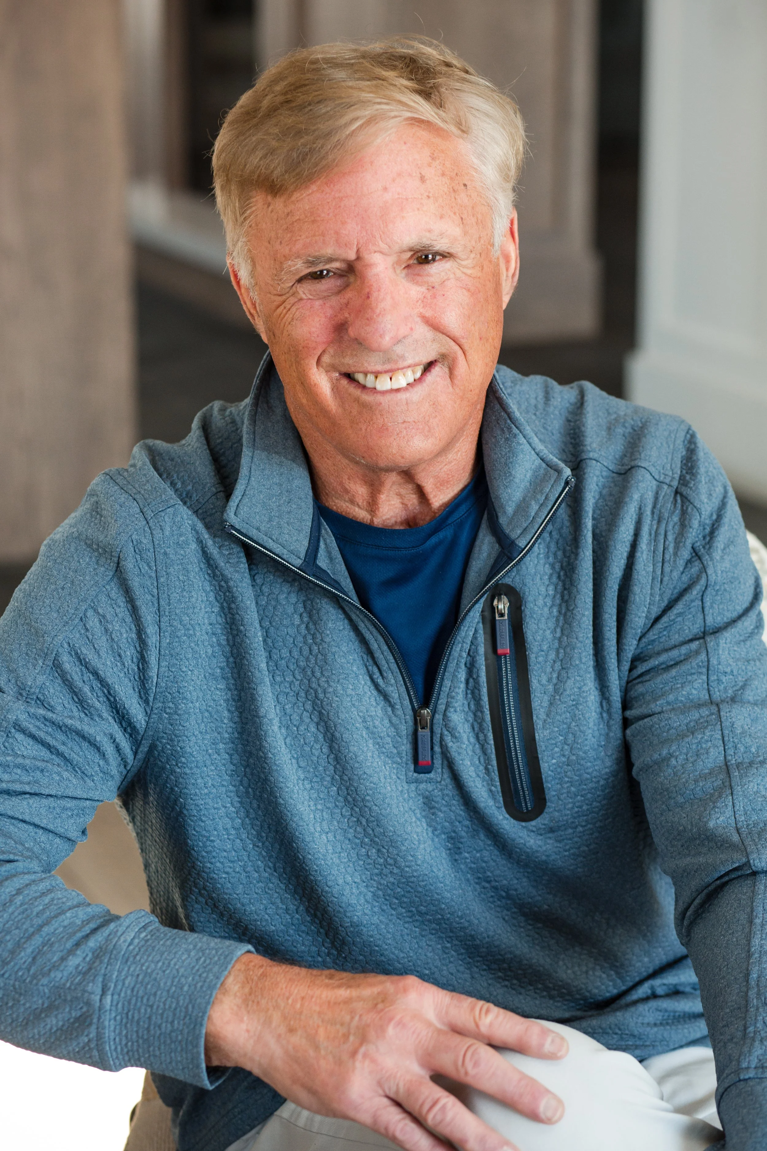 A smiling older man with light-colored hair wearing a blue zip-up jacket and a dark blue shirt, sitting indoors.