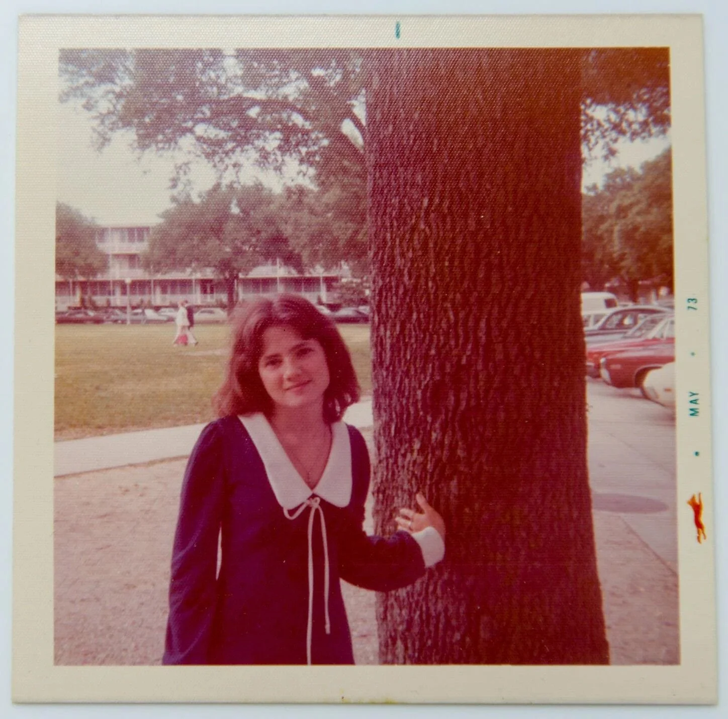 A Polaroid of Claudia Barker as a young girl, photographed in 1975, with brown hair, wearing a navy dress with a white collar, stands outdoors in New Orleans, next to a large tree.