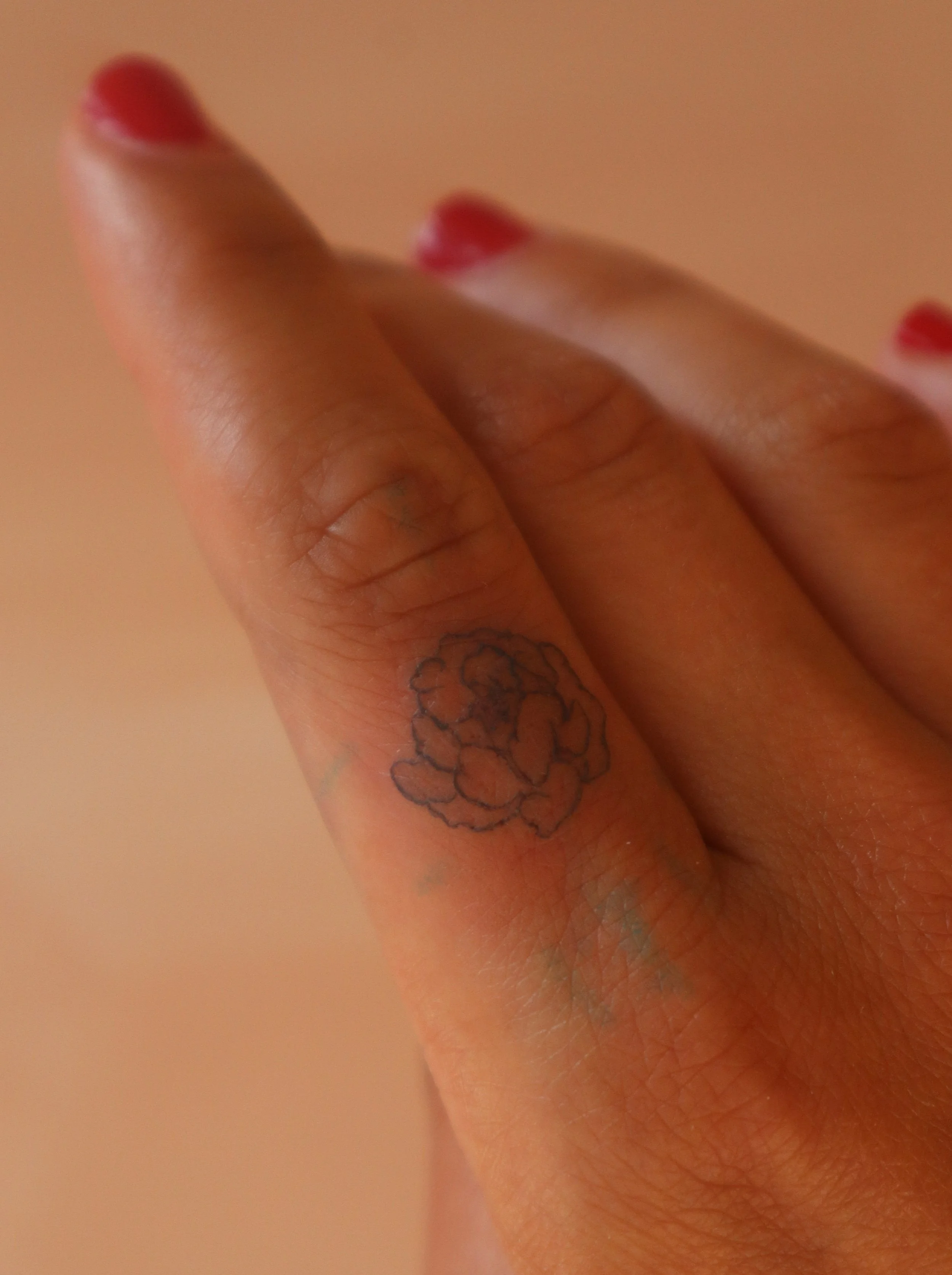 Close-up of a hand with red nail polish on the fingernails, showing a small flower tattoo on the middle finger.