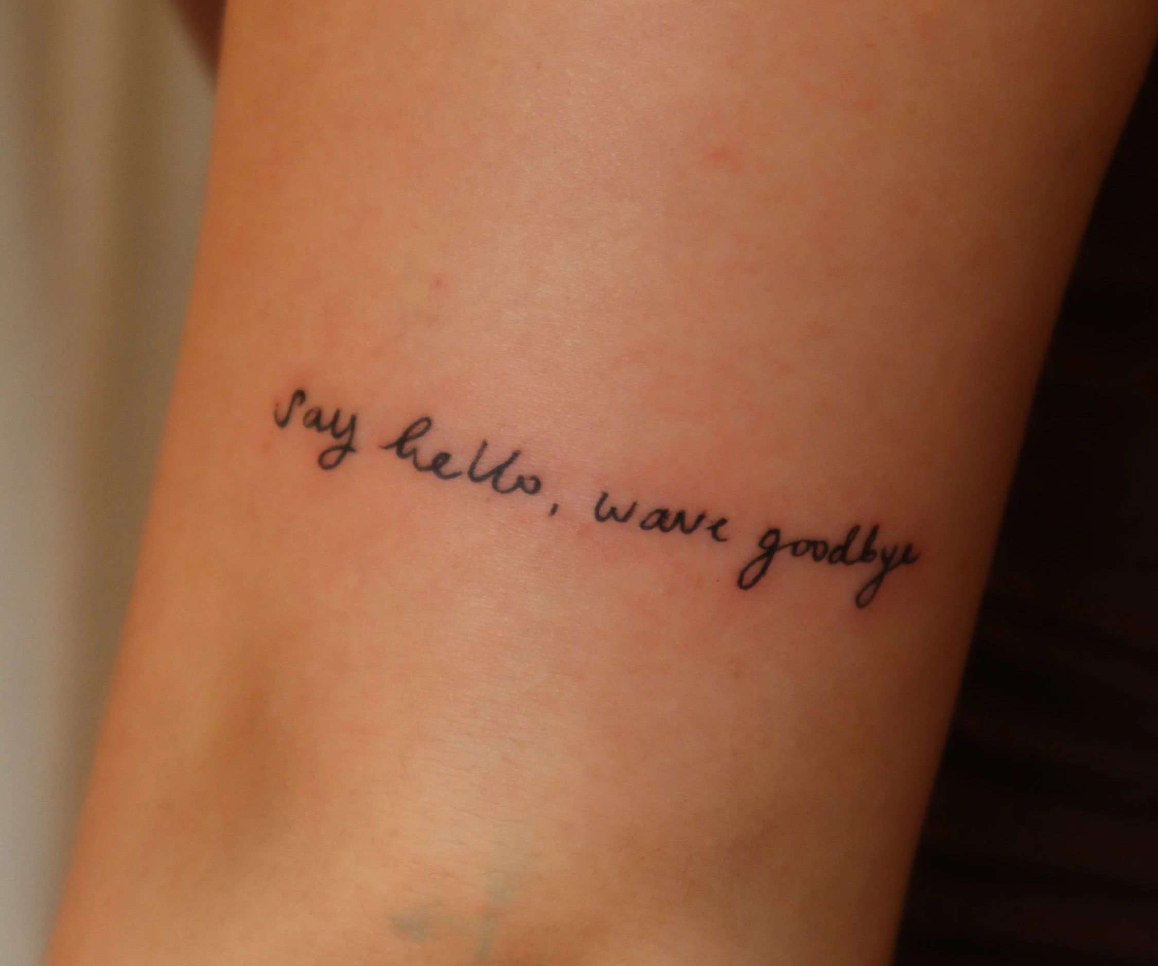 Tattoo on person's arm that says 'say hello, wave goodbye' in cursive font.