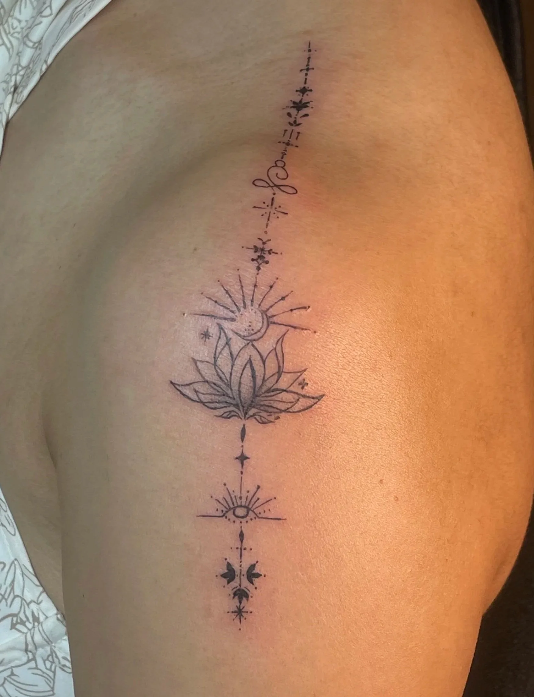 Tattoo on a person's side featuring a moon, sun, lotus flower, and celestial symbols arranged vertically.