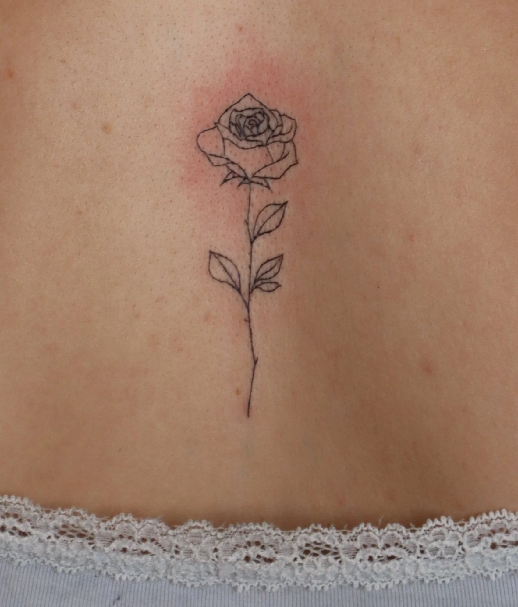 A delicate black ink tattoo of a single outline rose flower with leaves, located on the person's skin above a lace-trimmed fabric.