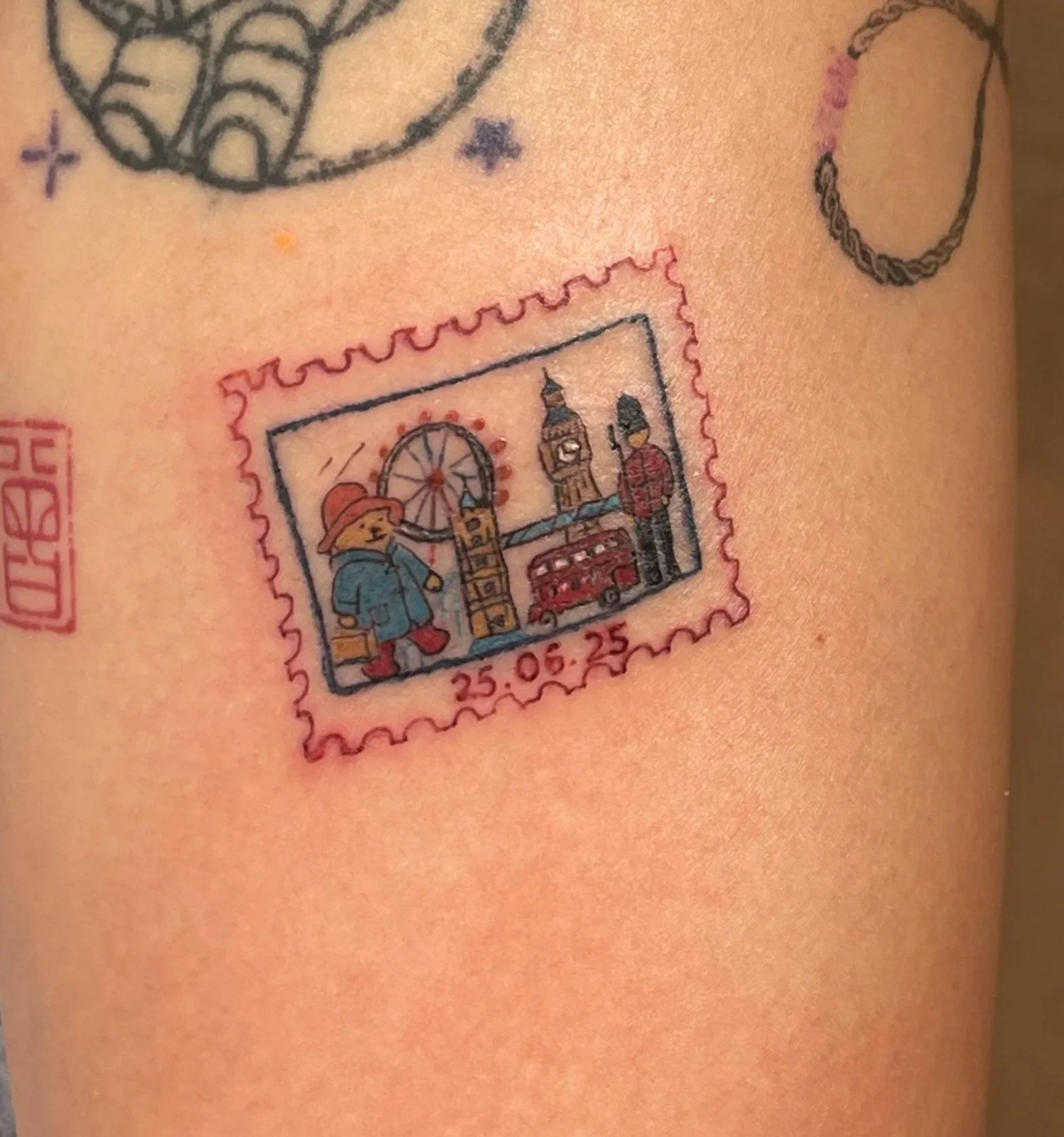 Tattoo of a stylized London scene on a person’s skin, featuring a bear in a hat and coat holding a bag, Big Ben, a red double-decker bus, a London guard, and a Ferris wheel, with the date 25.05.25 beneath the scene.