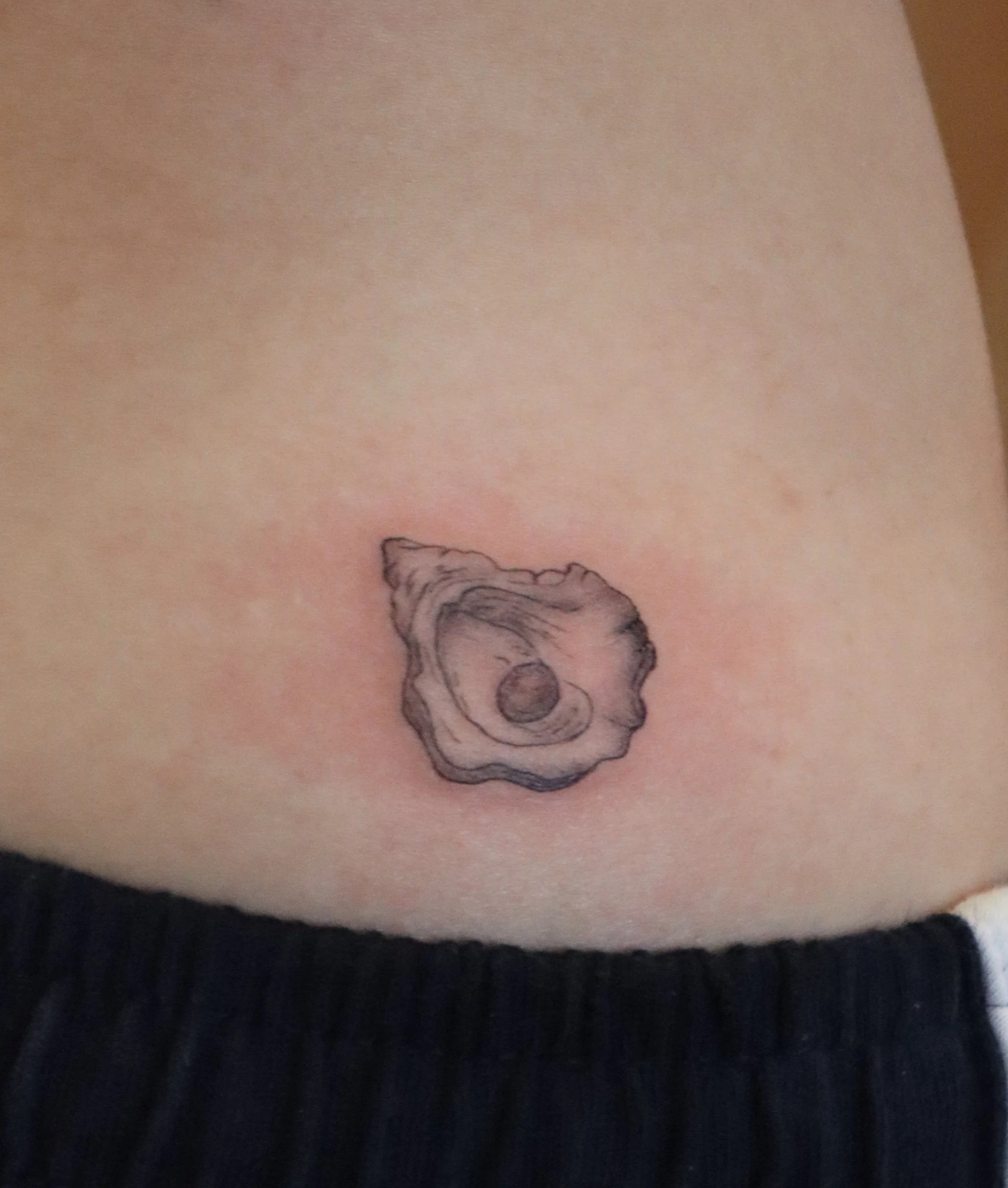 Tattoo of a seashell on the lower abdomen, above the waistband of black pants.