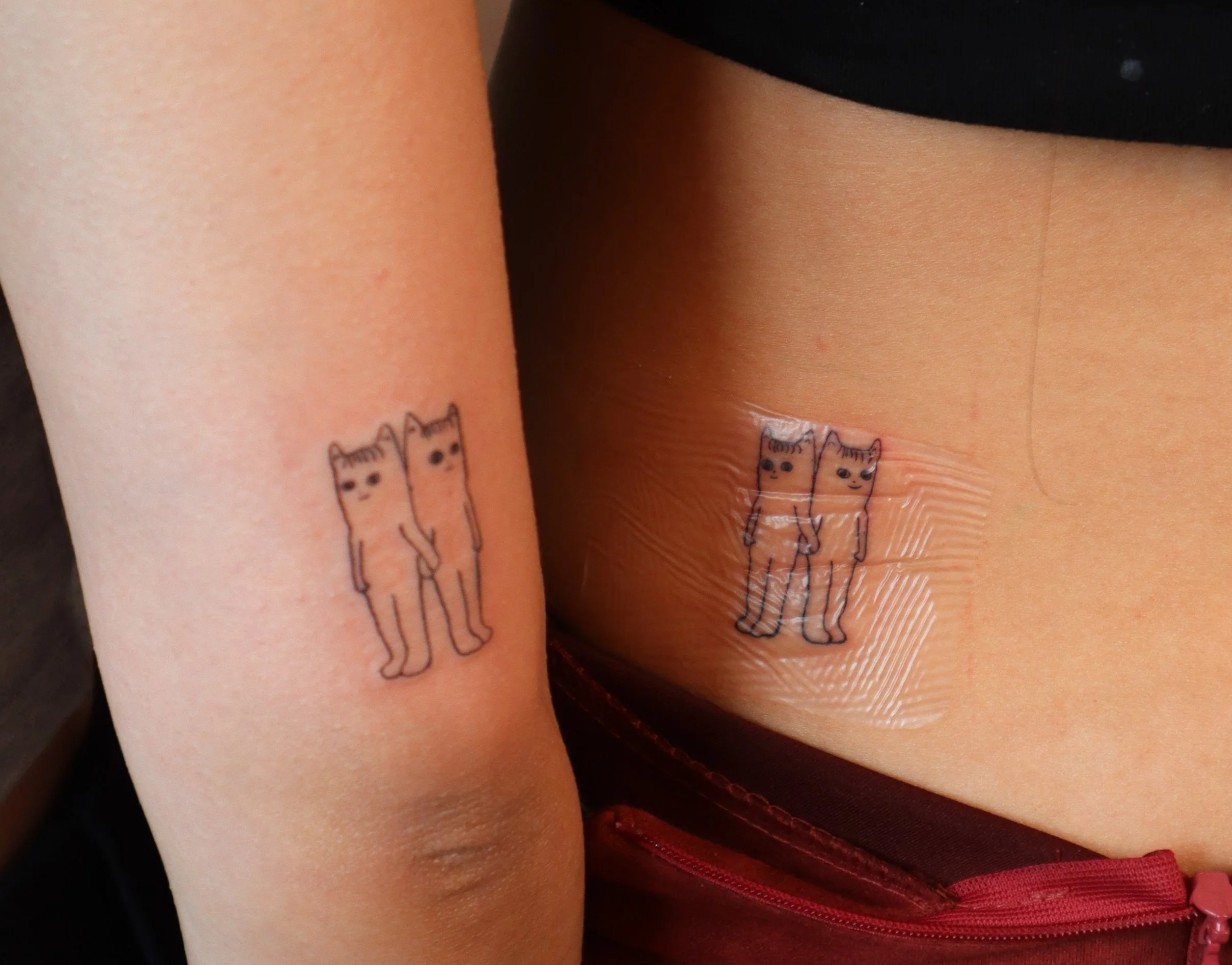 Two tattoos of cartoon cats on different parts of the body, one on the upper arm and one on the side of the torso, both showing two cats standing together.