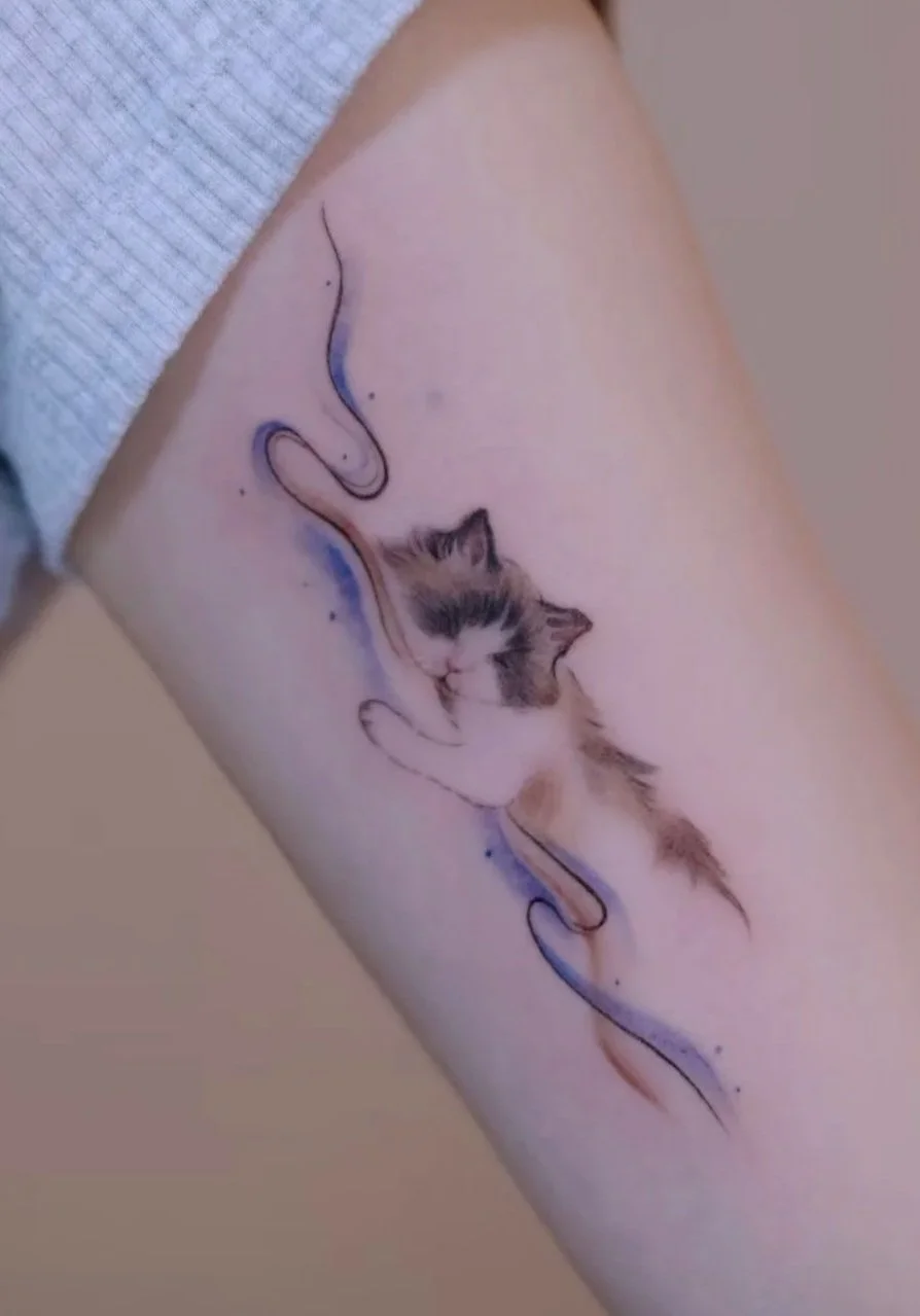 Tattoo of a sleeping kitten with a whiskered face and closed eyes, being gently snuggled by a stylized line drawing of a hand, on a person's arm.