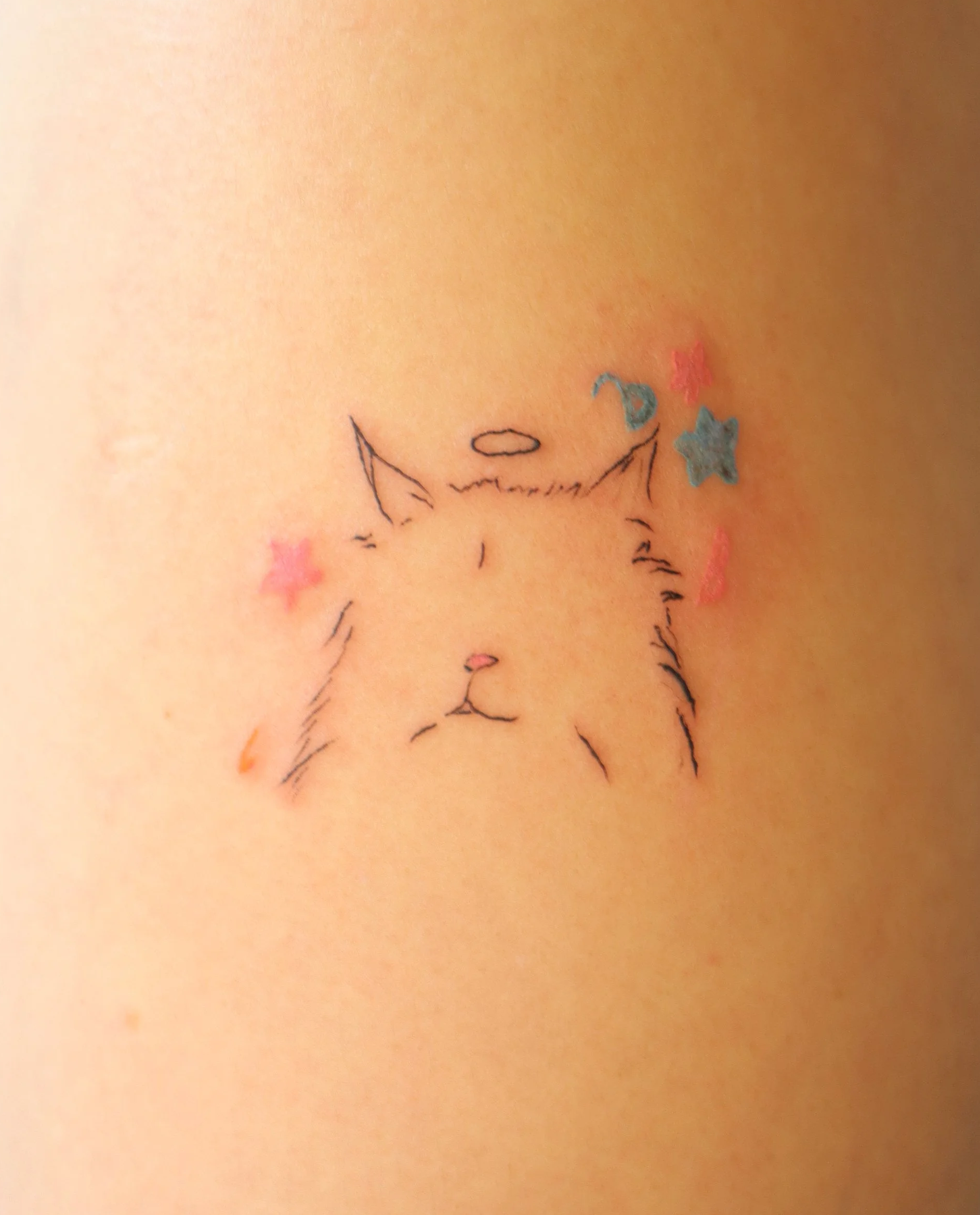 Tattoo of a simple line drawing of a cat with a small halo above its head, surrounded by colorful star and moon stickers.