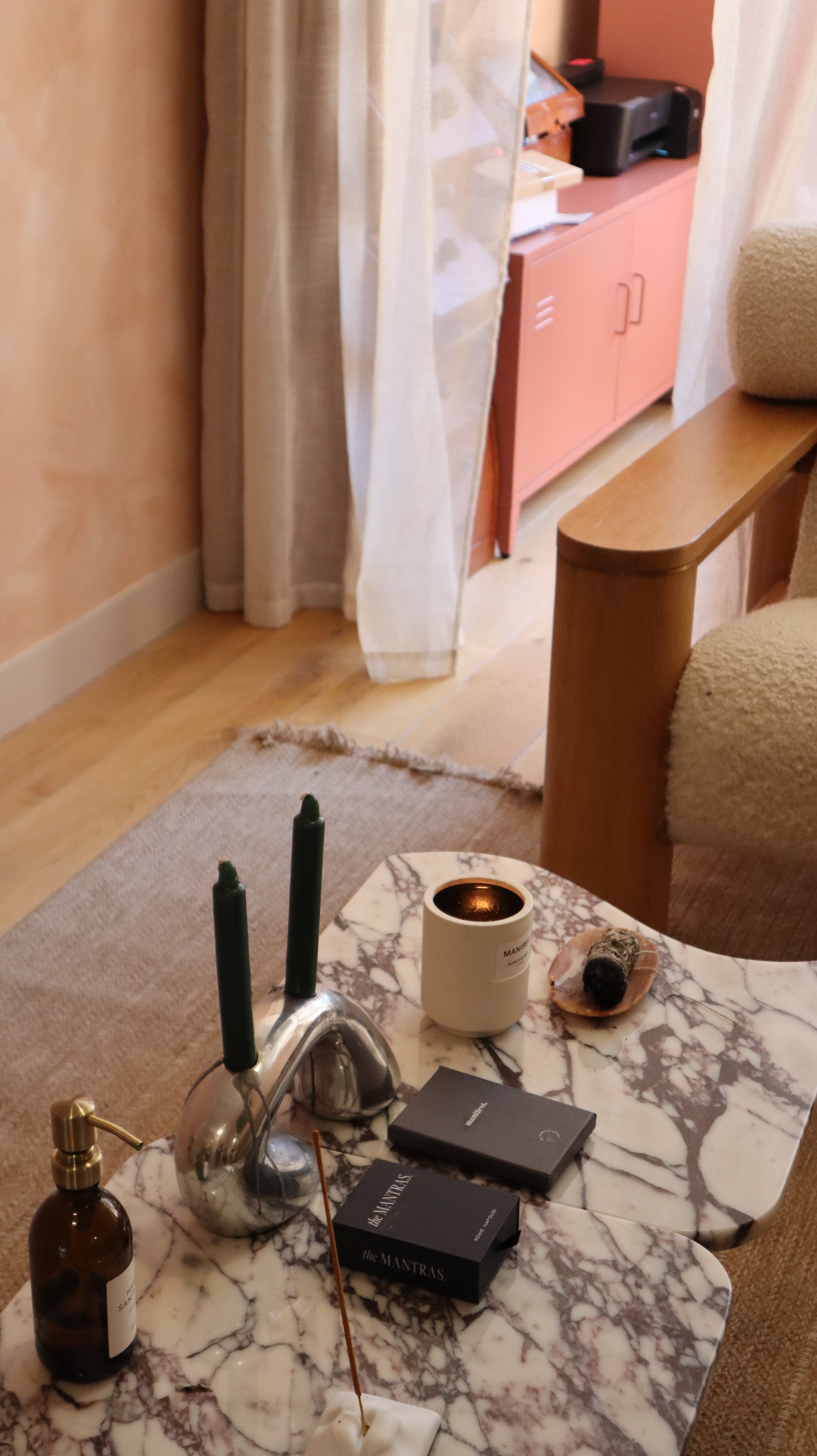 Close-up of a marble coffee table with dark green candles, a brown candle holder, a small white candle, a rock, and black boxes labeled 'the MANTRAS'. In the background, a beige armchair partially visible, with a pink cabinet and a black printer in the corner. There is a curtain and a carpet on the floor.