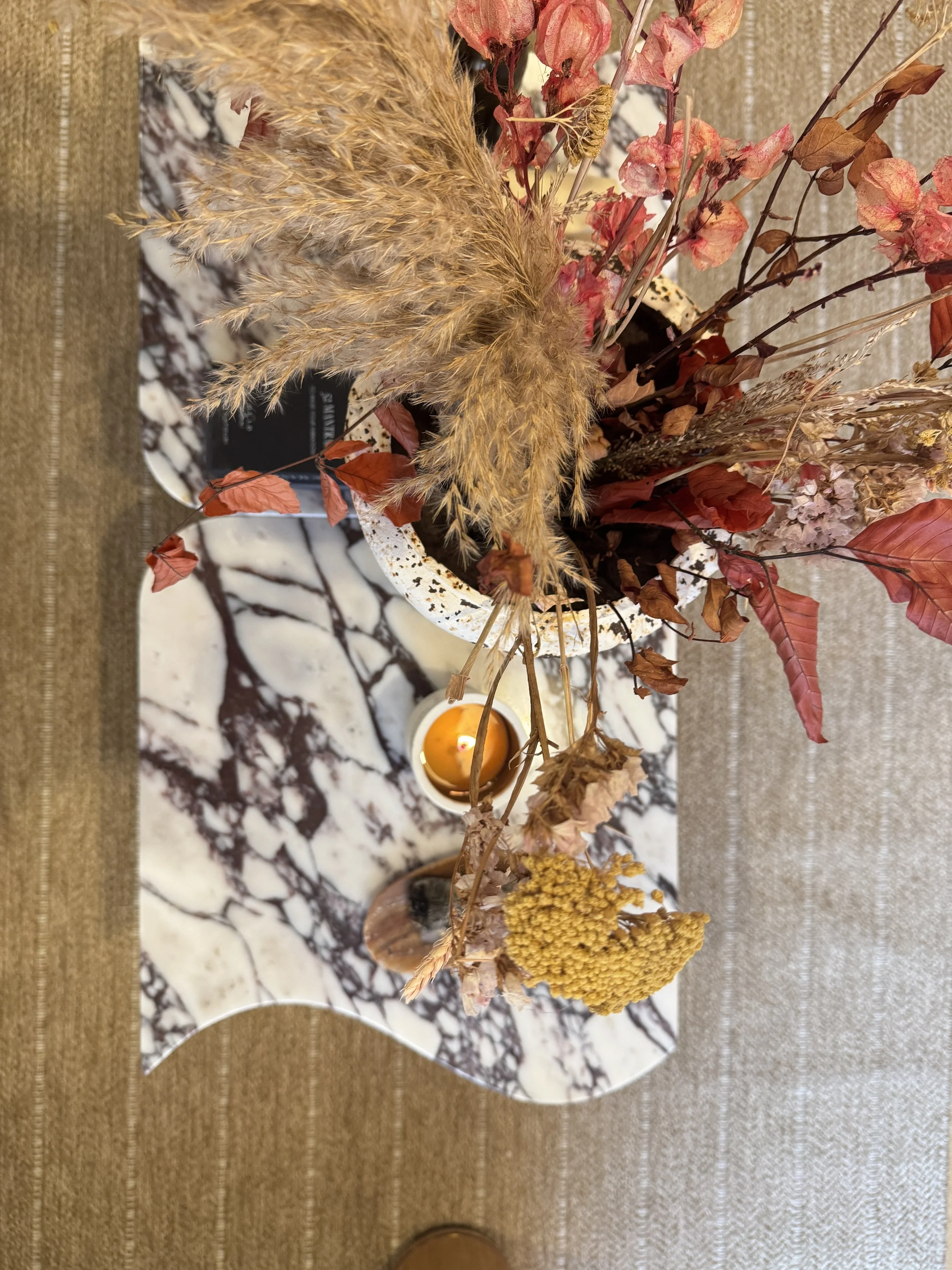 A floral arrangement in a decorative vase featuring dried flowers and grasses, with a small candle in a holder nearby. The arrangement includes pink, red, and yellow dried flowers, and the setup is on a marble and wood surface.
