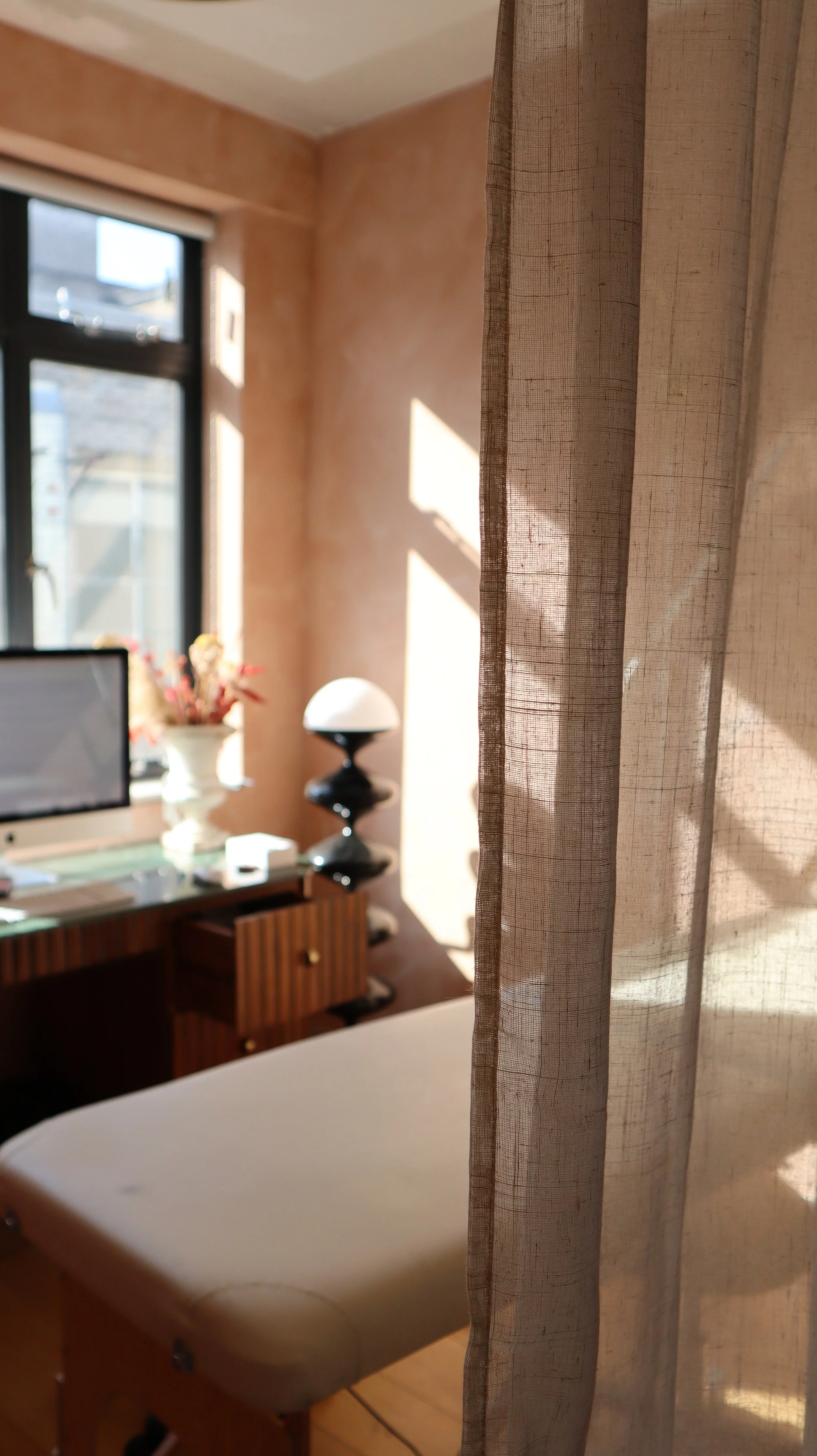 A beige bathtub seen in front of a semi-transparent curtain, with a wooden floor. In the background, a wall with a window, a side table with a lamp, a computer, and a vase with flowers are visible.