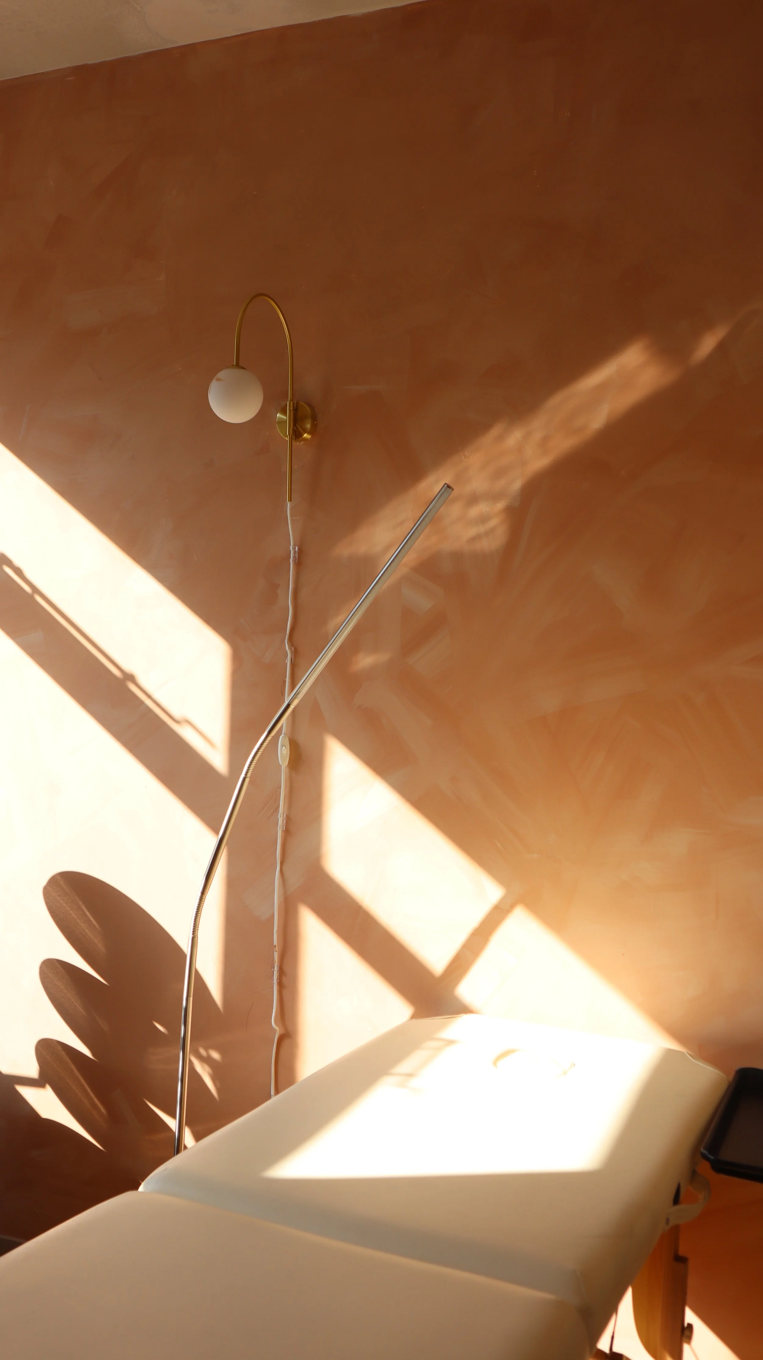 A hospital or clinic examination room with a beige examination table covered in a white sheet, a wall-mounted lamp with a white globe shade, and a shadow of a plant on the wall. Sunlight streams into the room.