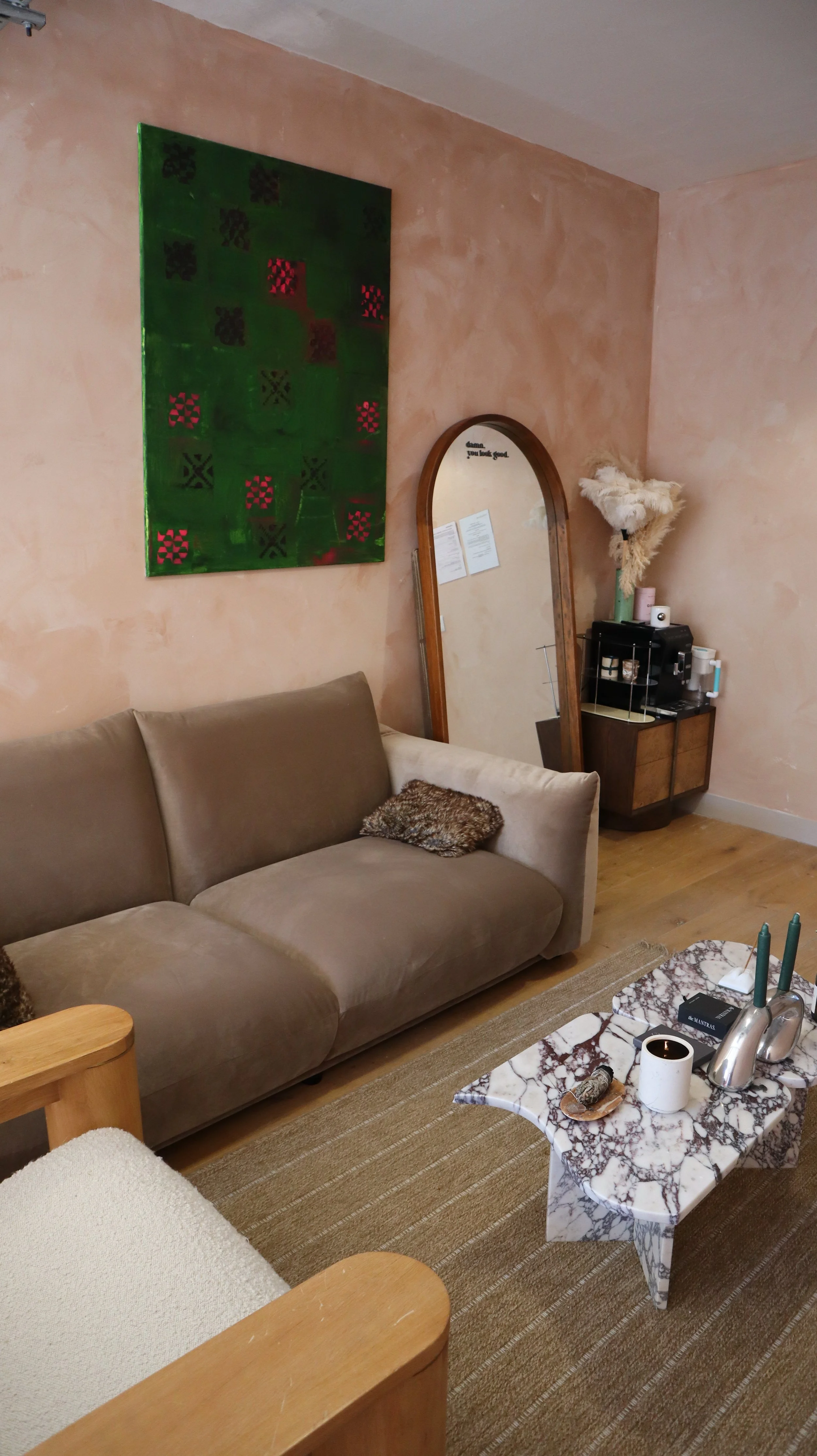 A living room corner with a beige couch, a small leopard print pillow, a white marble coffee table with candles and decor, a tall mirror, a textured pink wall with abstract art, and a corner shelf with plants and objects.