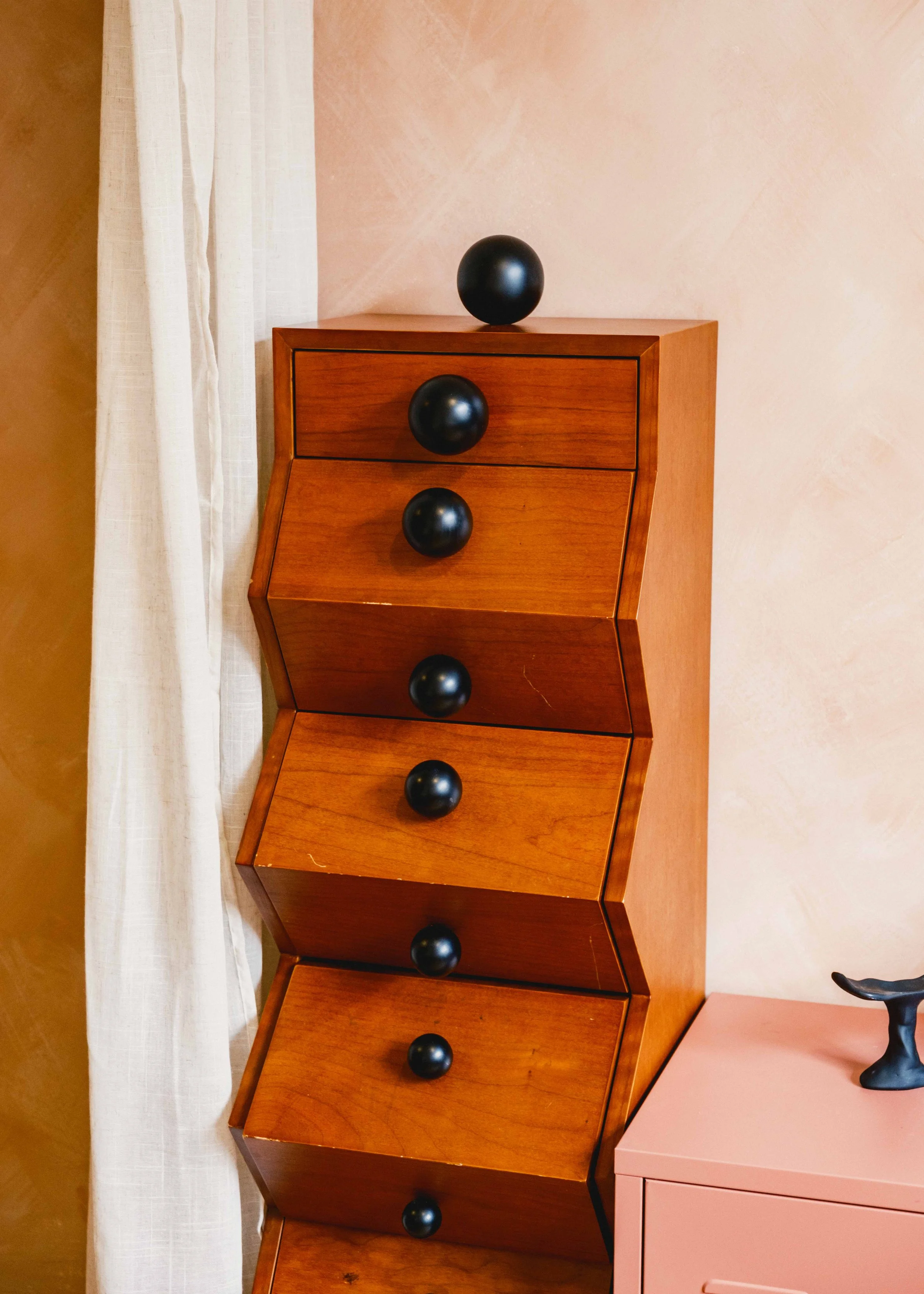 Wooden cabinet with black spherical knobs, positioned next to a white curtain and a pink dresser with a black decorative object on top, against a pink wall.