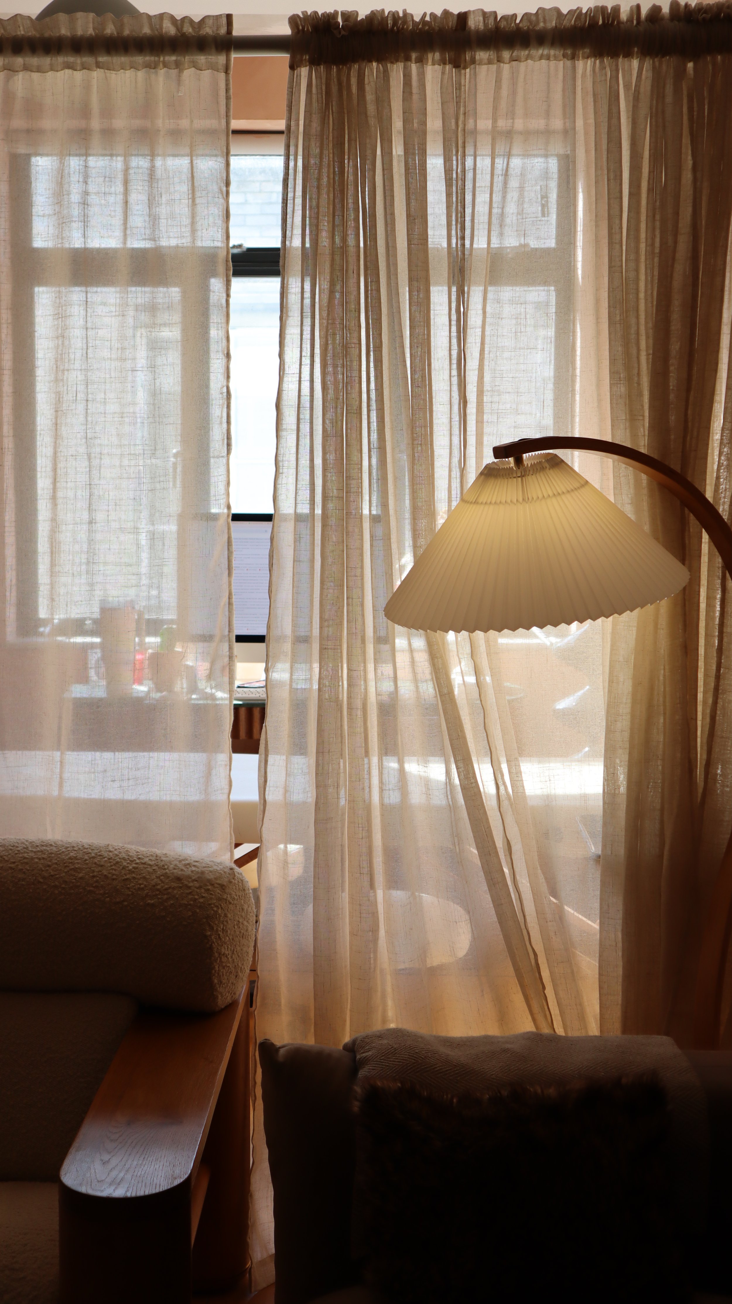 Living room view through sheer curtains showing a window and a lamp with a beige shade.