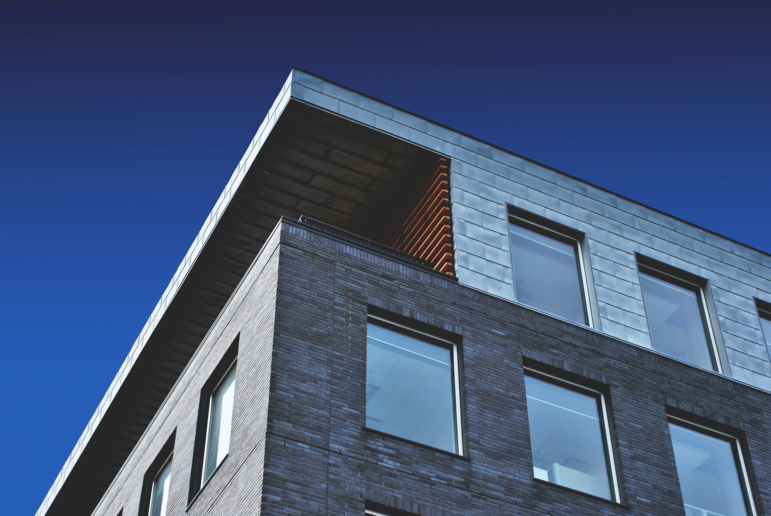 Modern multi-story building with dark brick exterior, large rectangular windows, and a metal facade on the top level, against a clear blue sky.