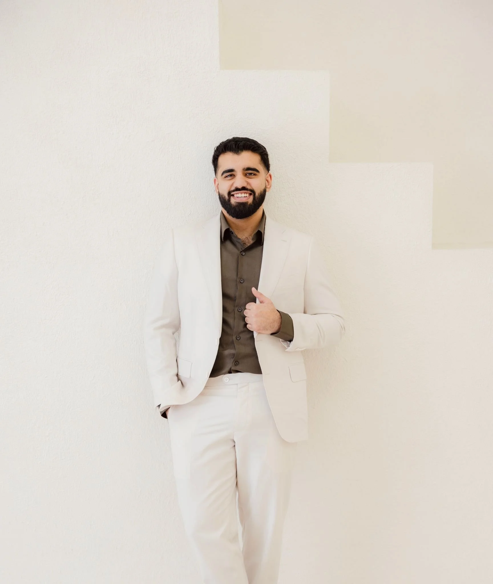 A man with dark hair and beard wearing a beige suit and brown shirt, smiling and posing against a plain light-colored wall.