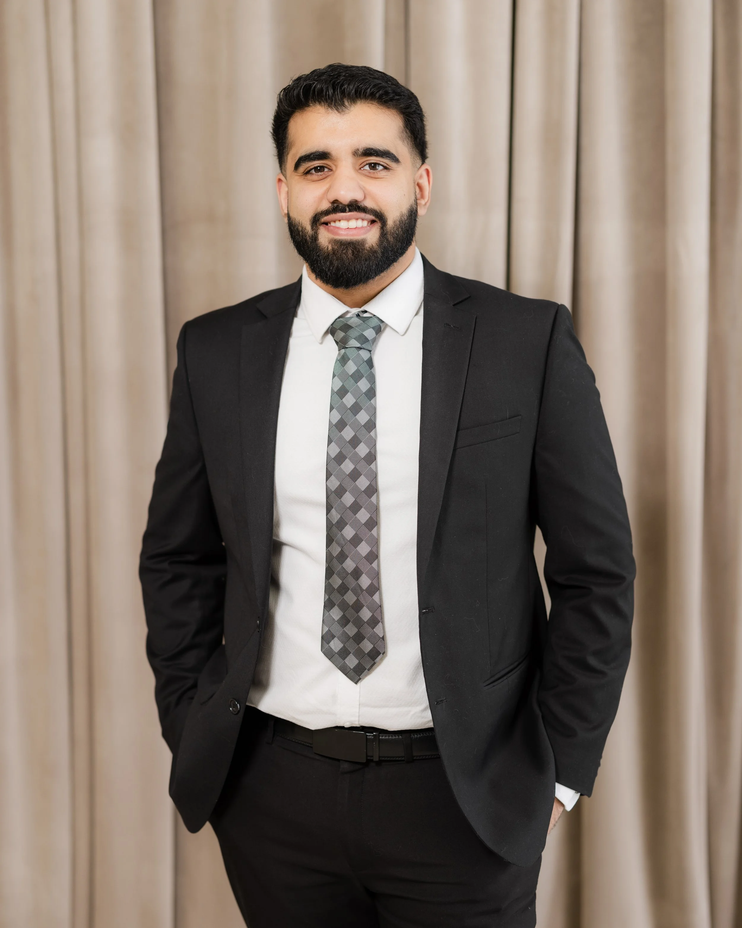 A smiling man in a black suit, white shirt, and a patterned gray tie standing in front of beige curtains.