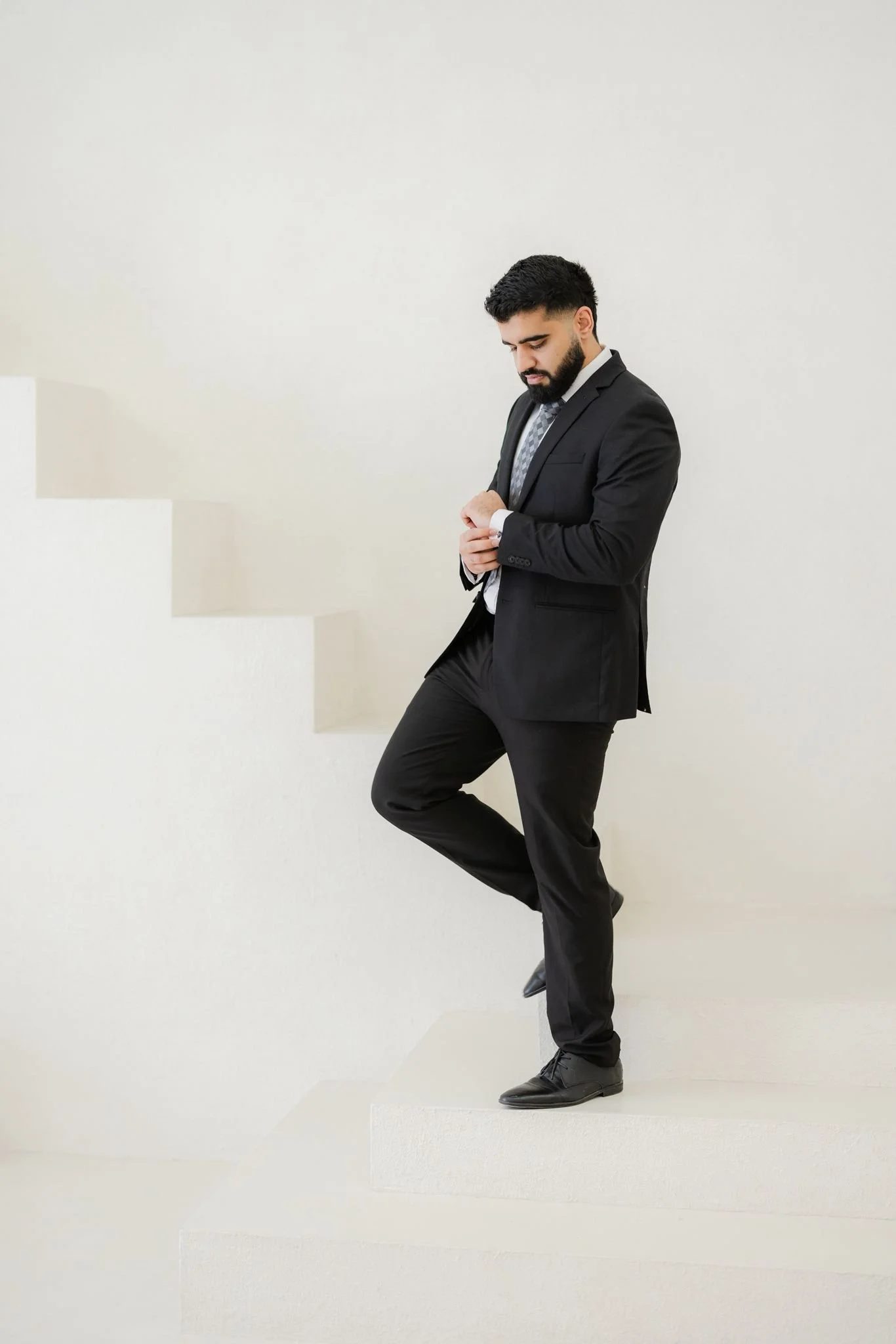A man in a black suit and tie standing on stairs with a plain light-colored background.