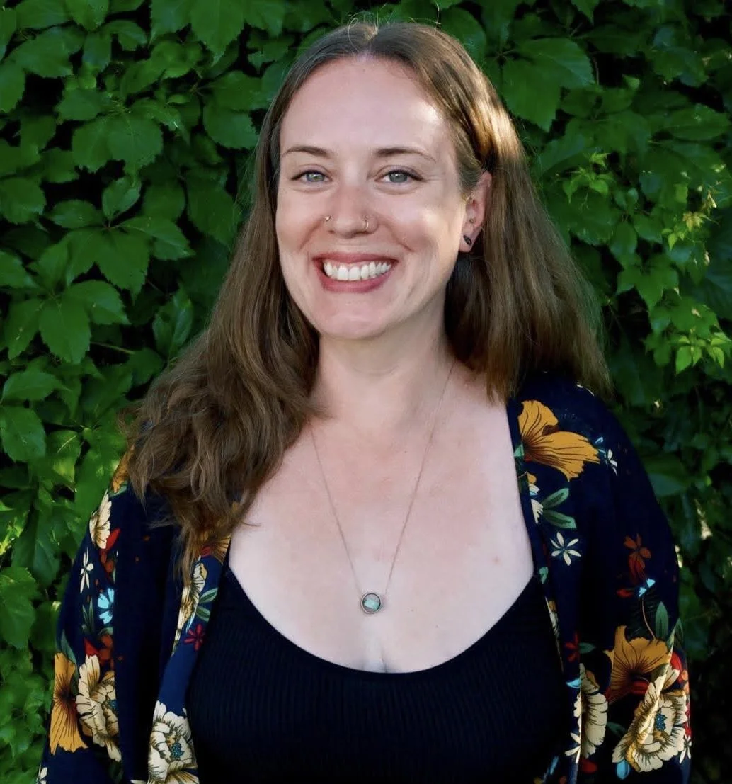 A woman with long brown hair and blue eyes smiling outdoors in front of green leafy plants, wearing a dark floral top.