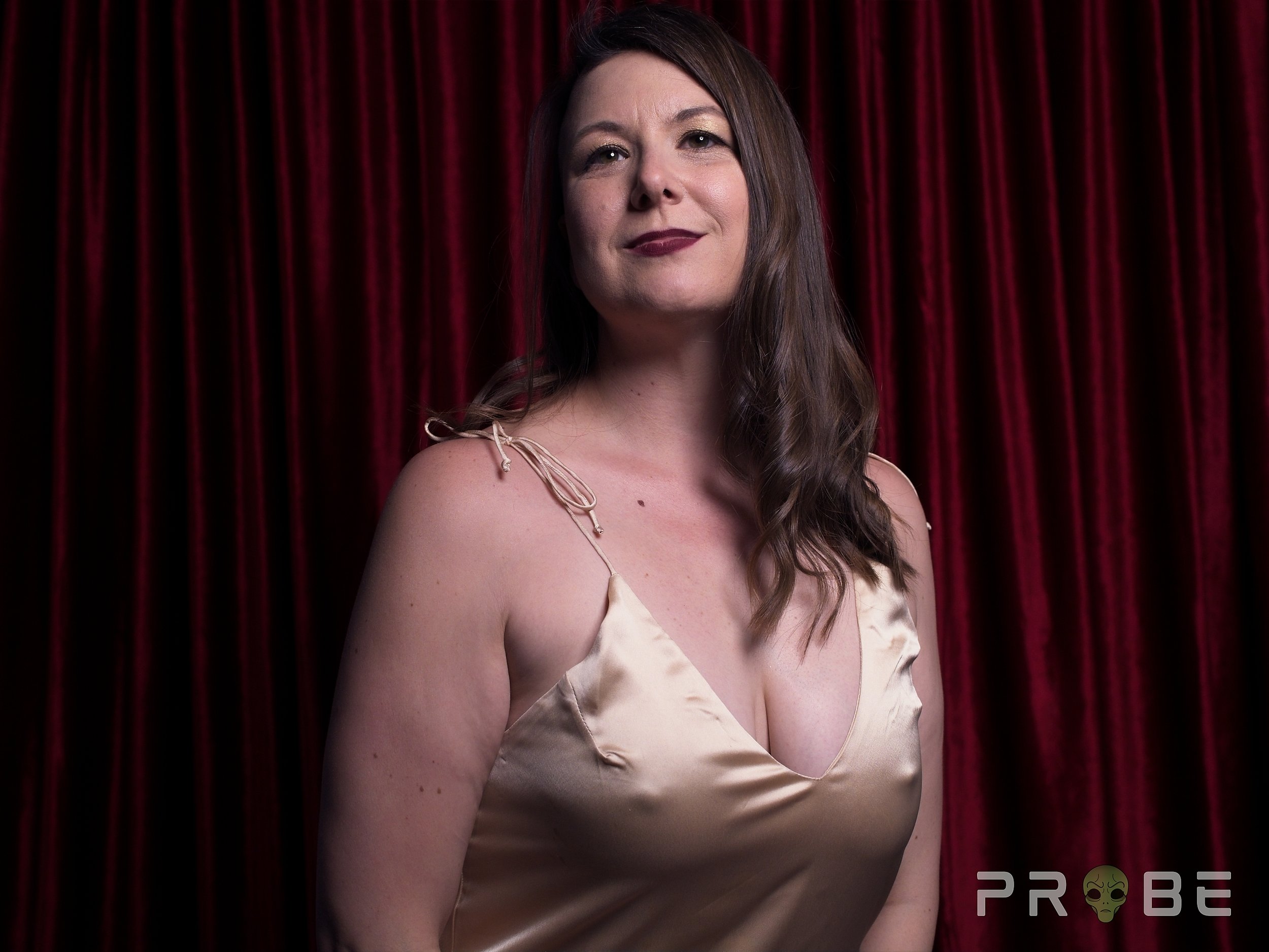 A woman with long brown wavy hair wearing a light-colored satin dress, standing in front of a red curtain.