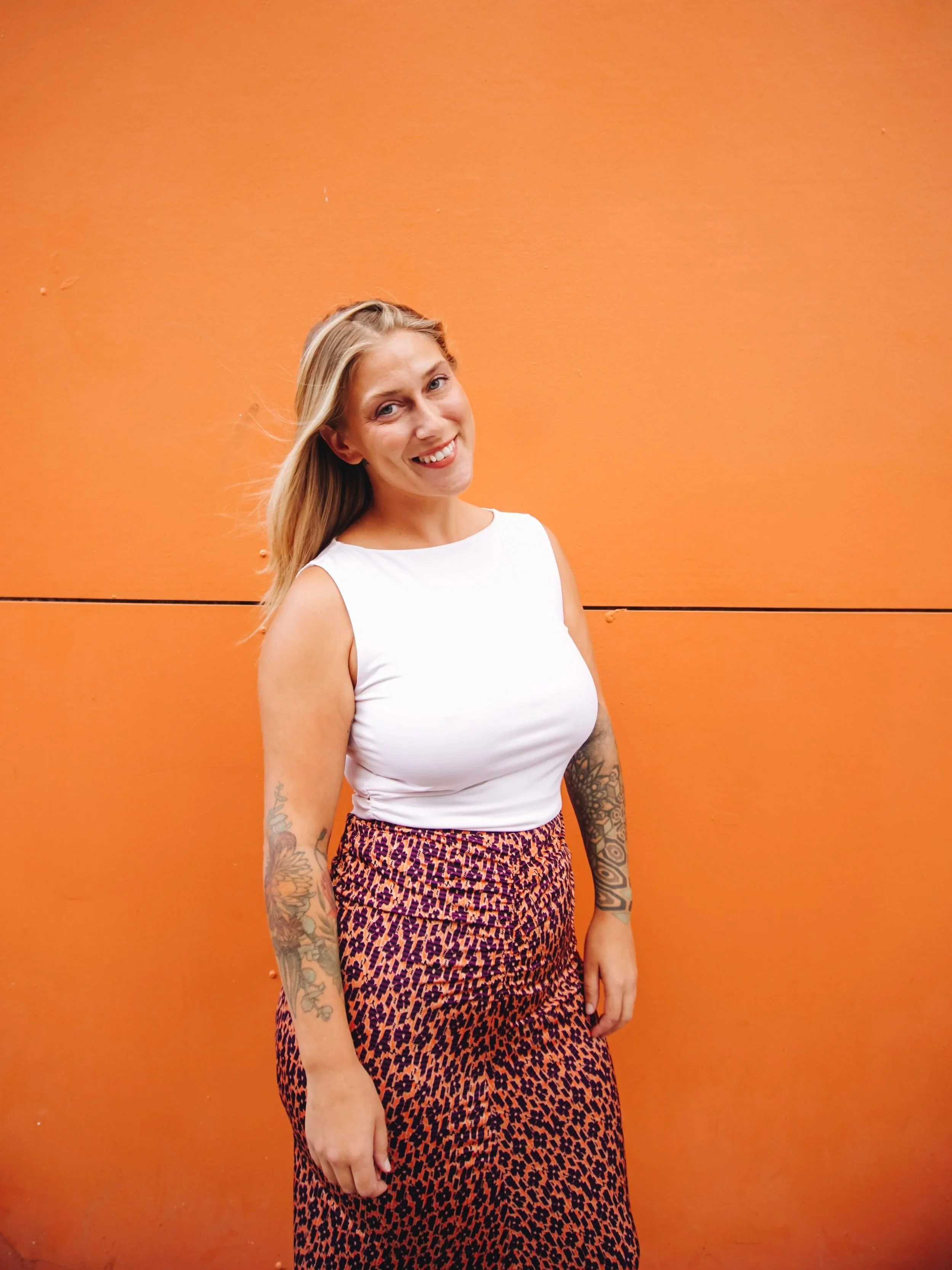 A woman with shoulder-length blonde hair, smiling, wearing a sheer long-sleeve top with colorful abstract patterns and high-waisted blue shorts, standing against a beige background.