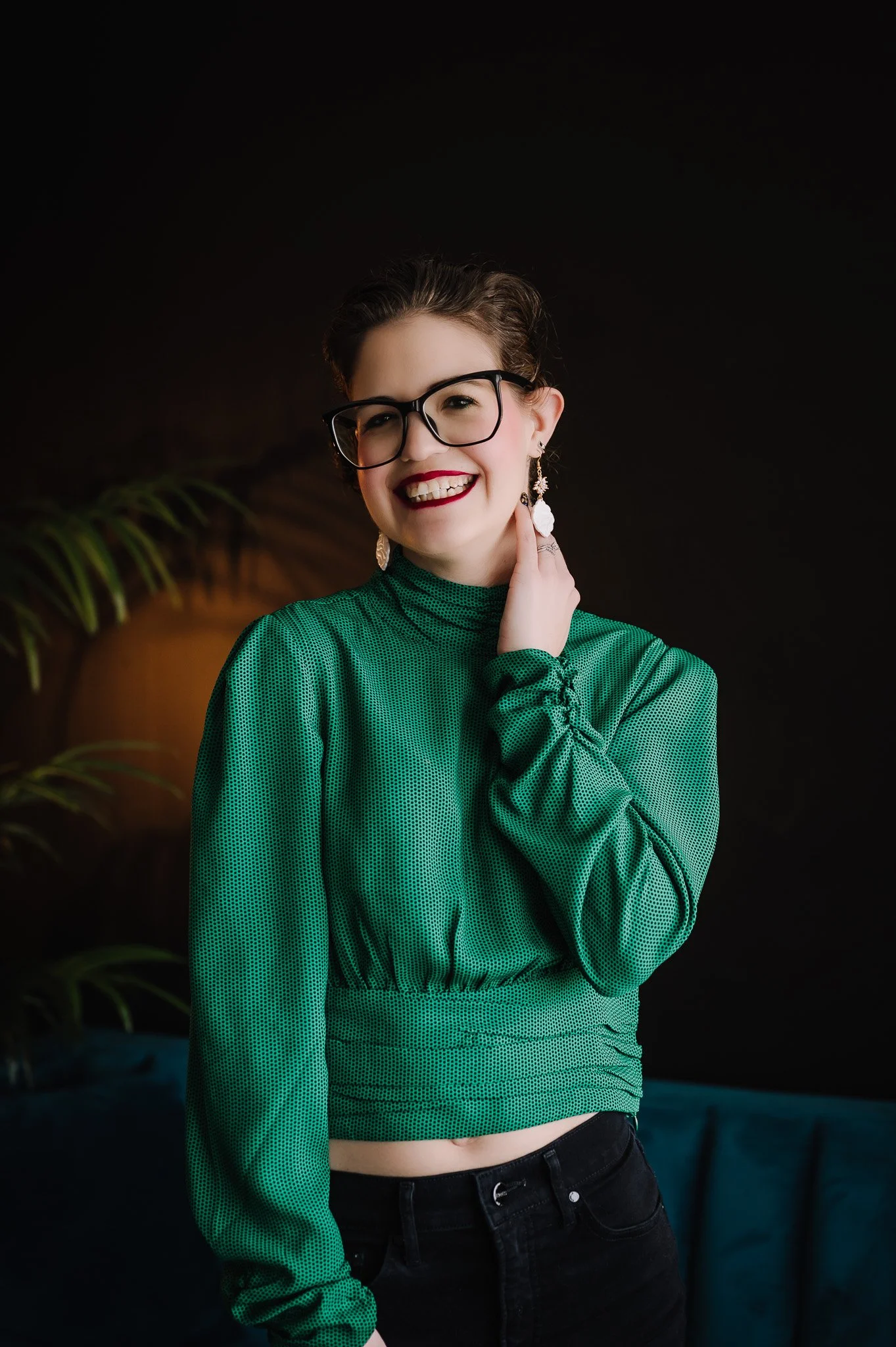 Young woman with glasses, earrings, and a green top smiling against a dark background.