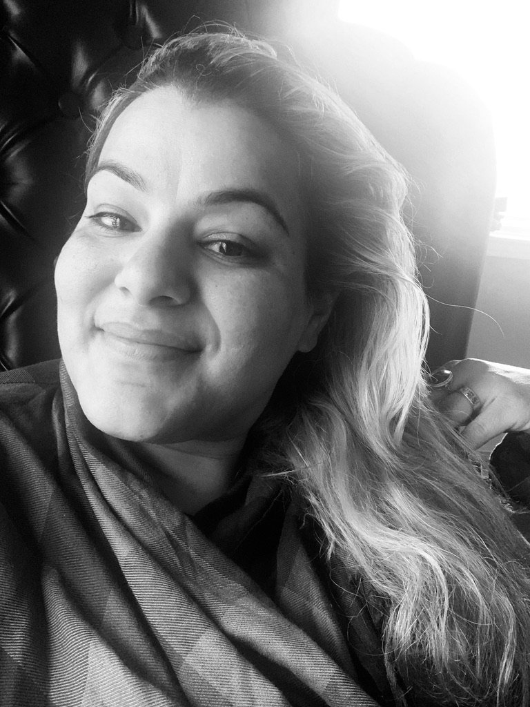 Black and white photo of a smiling woman with wavy hair, sitting on a leather chair, with sunlight coming through a window in the background.